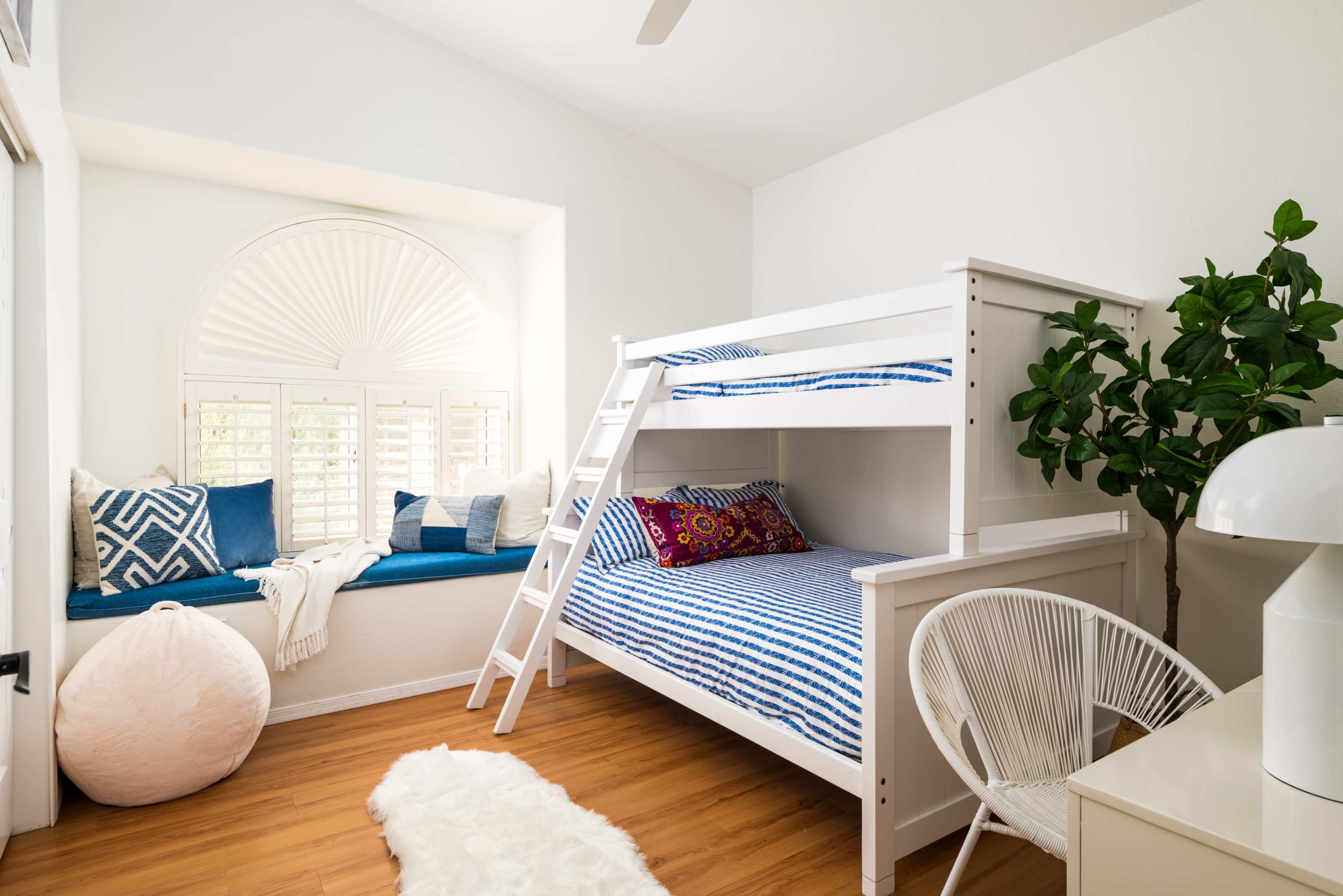 A bright bedroom features a bunk bed, a cozy seating area by a large window, and a plant in the corner.