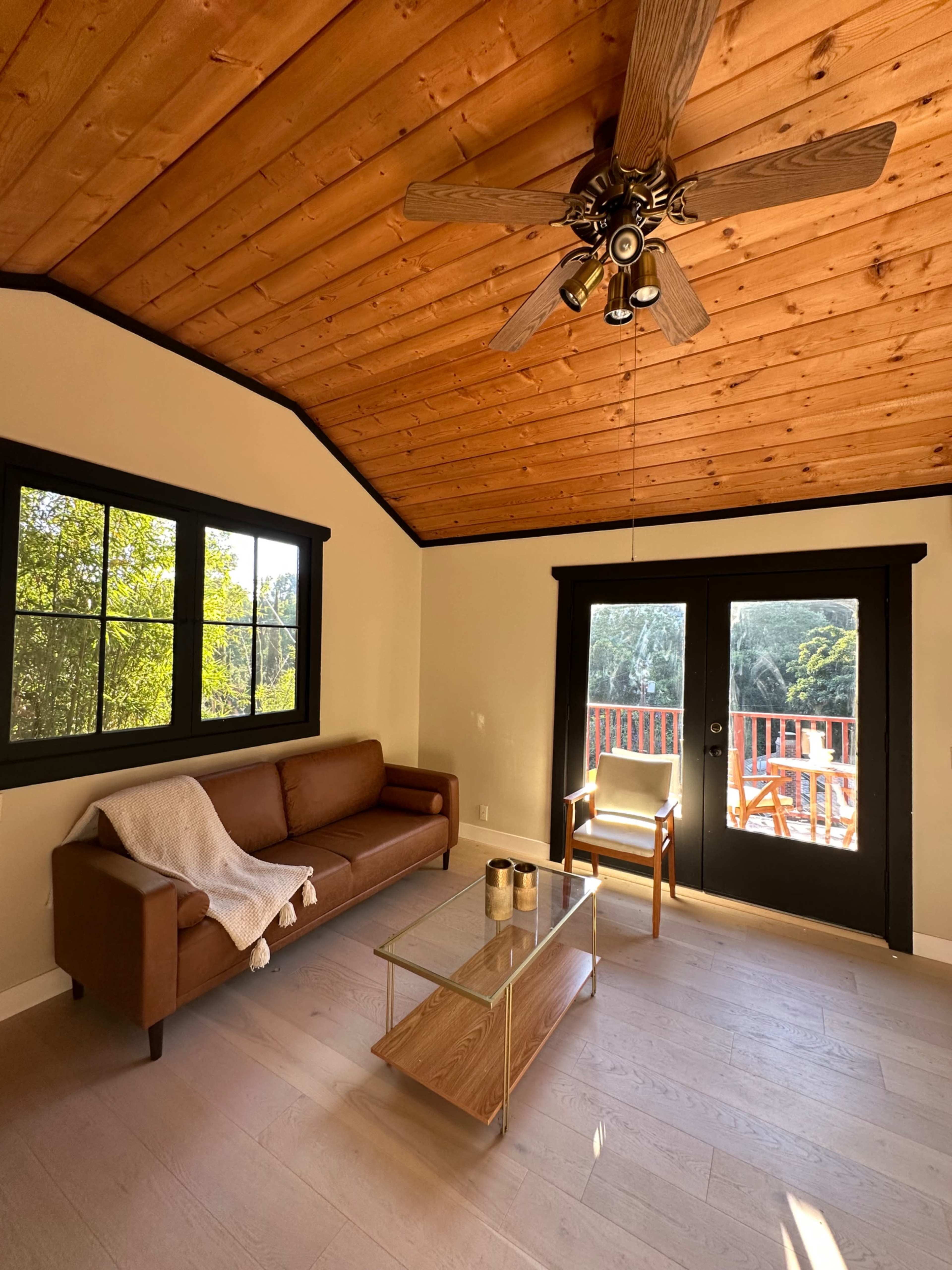 A cozy living room features a brown sofa, a glass coffee table, and a chair, with a wooden ceiling and large windows overlooking a deck.