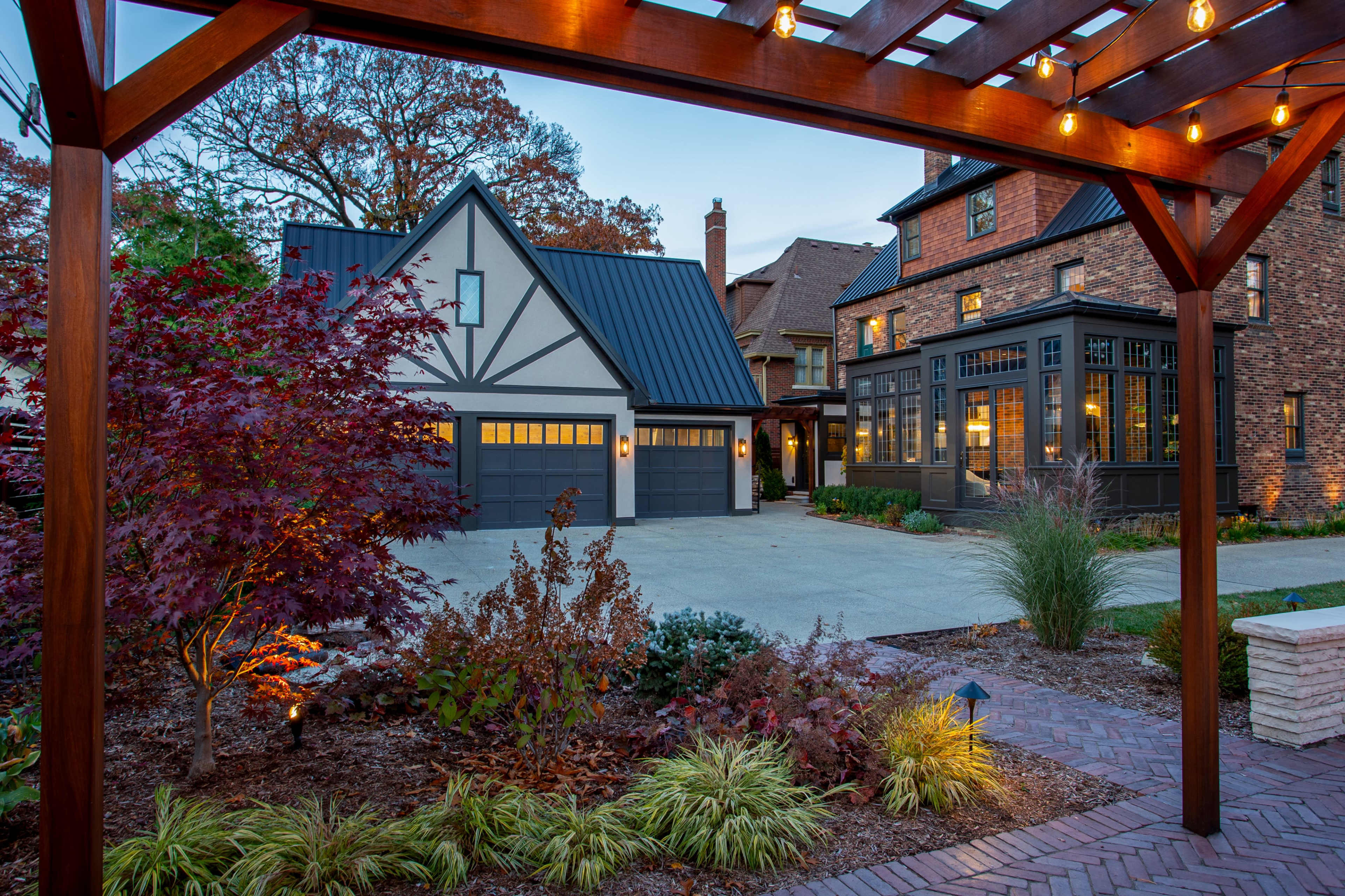 A landscaped residential area featuring two distinct houses, a garage with a modern roof, and outdoor lighting under a wooden pergola.