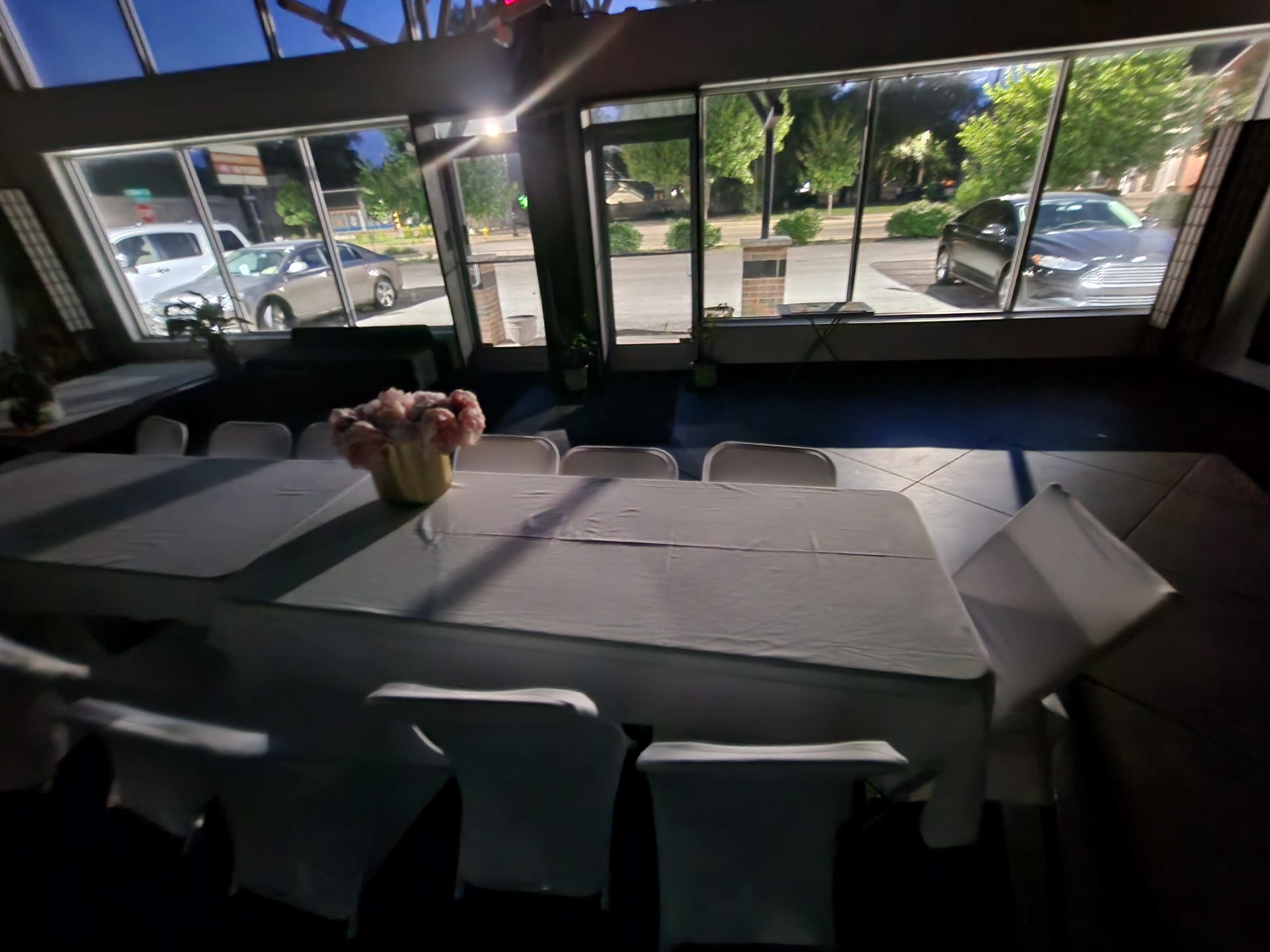 A long table covered with a white cloth is set in a room with large windows overlooking a parking lot and trees outside.
