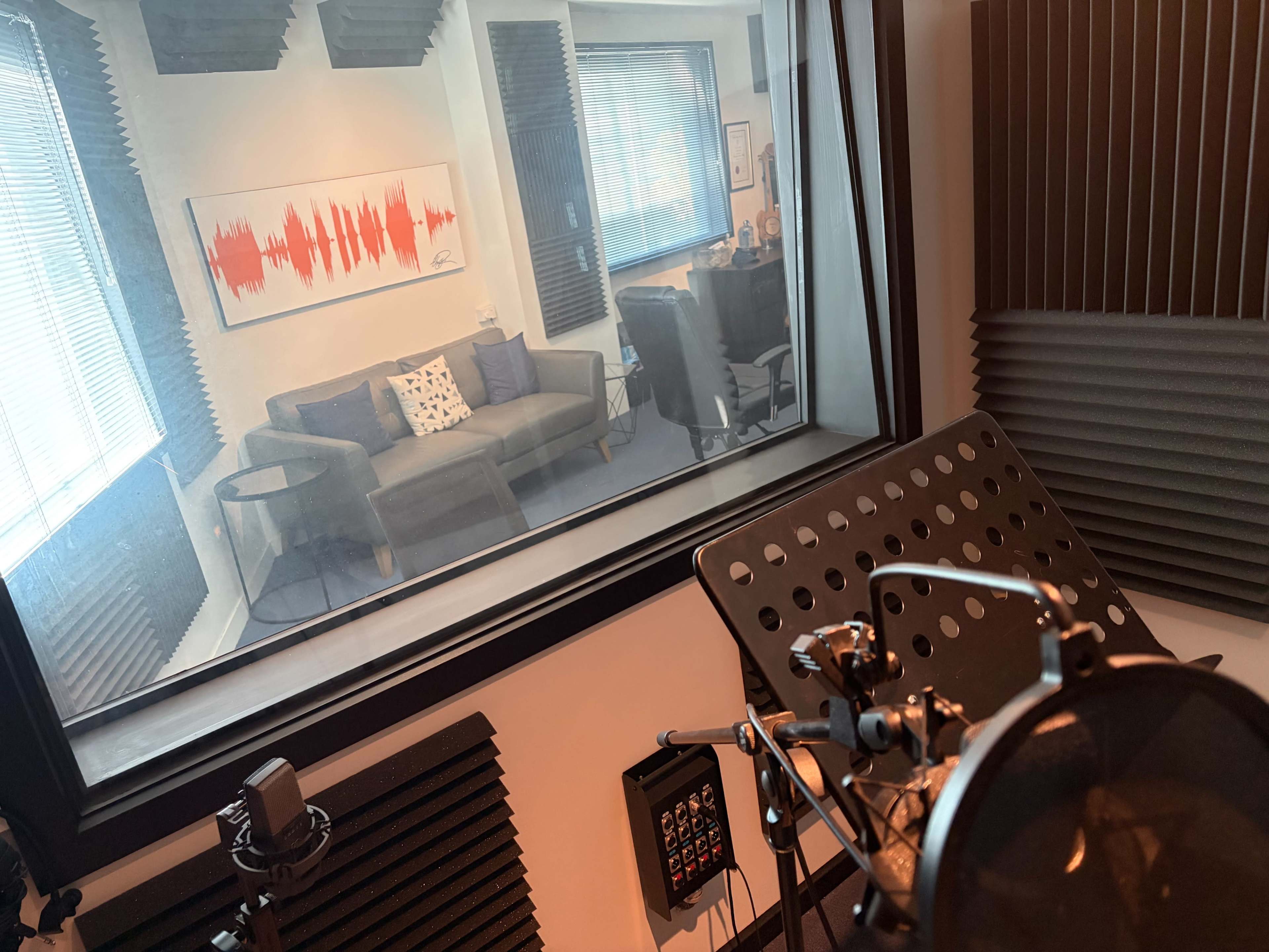 The image shows a soundproof recording booth with a microphone and music stand in the foreground, and a visible studio space containing a couch and armchair in the background.