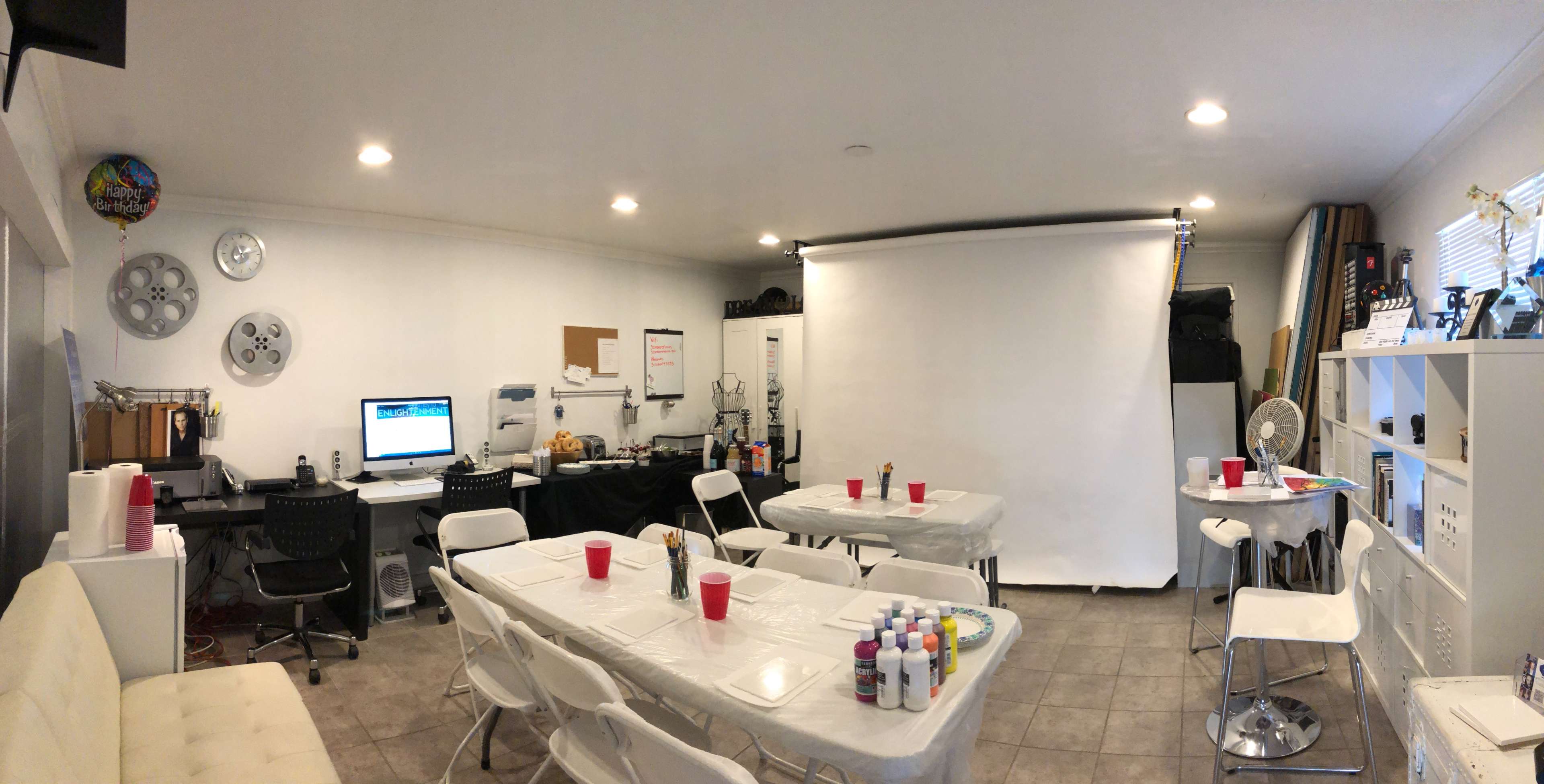 An art studio set up with tables covered in white cloth, red cups, and paint supplies, alongside a large blank backdrop for presentations or activities.