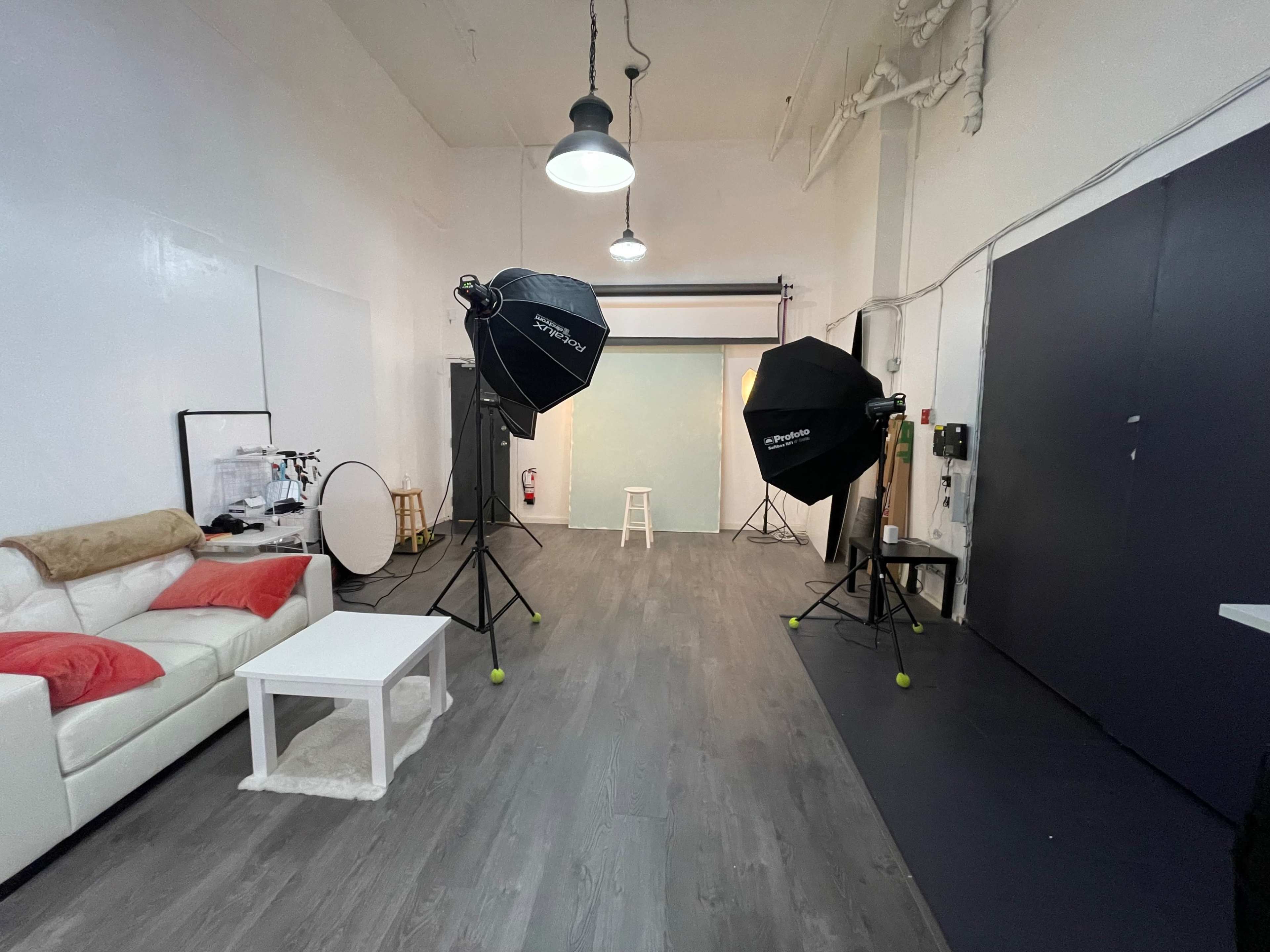 A photography studio features a white wall, wooden flooring, a white couch with red pillows, studio lights, and a backdrop setup.
