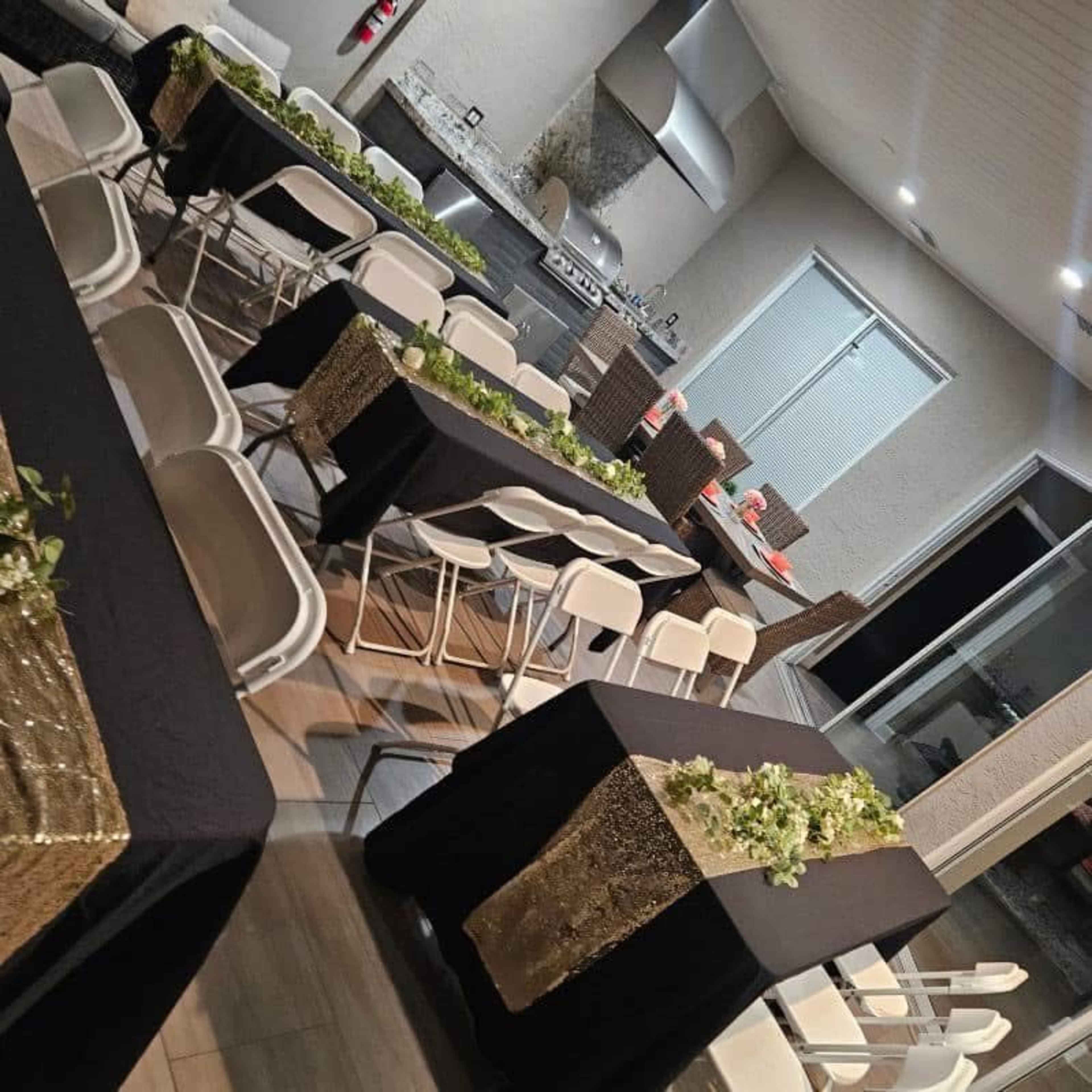 The image shows a dining area set up with black tables and white chairs, decorated with greenery and gold accents, and featuring a grill in the background.