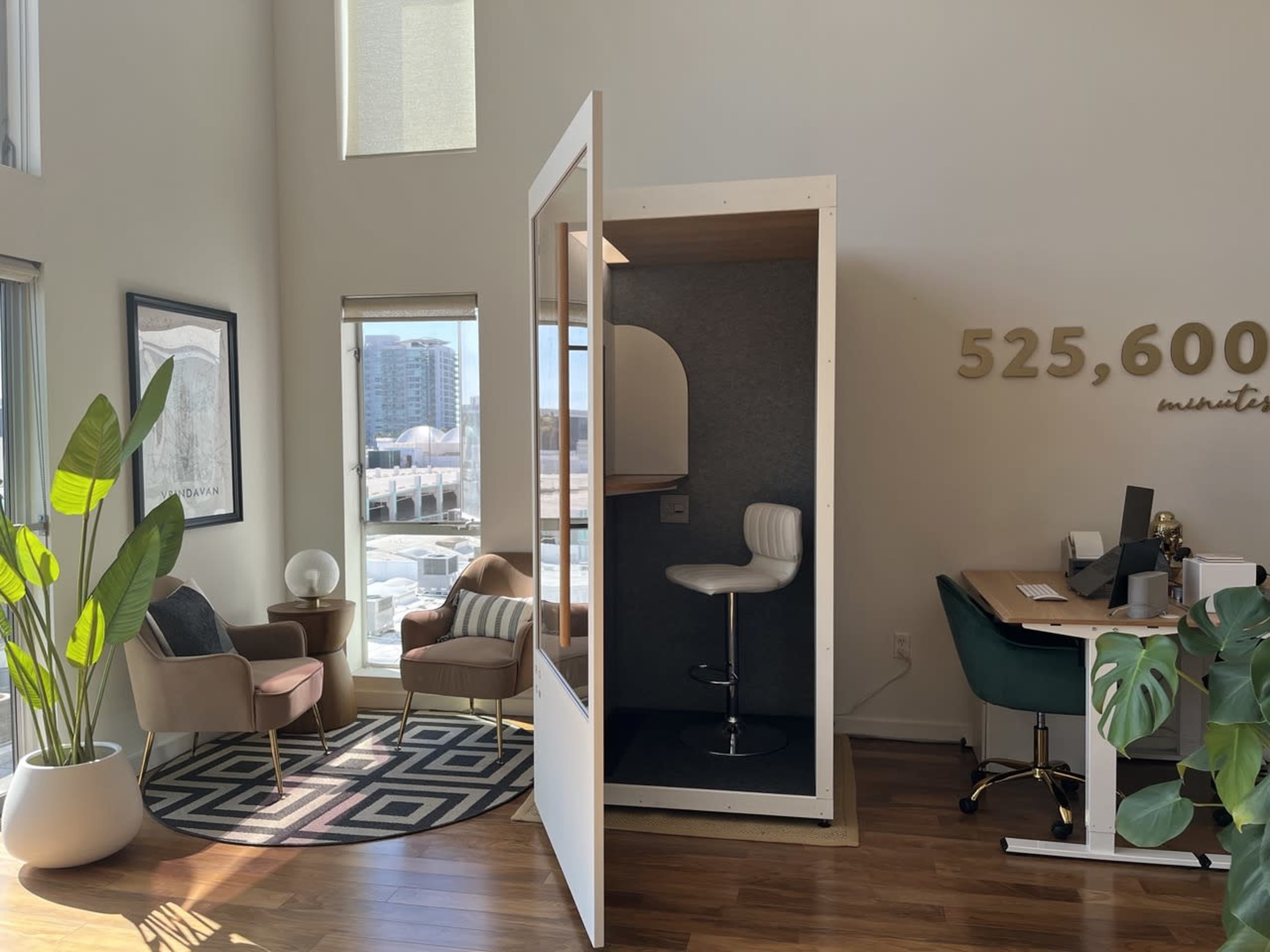 A modern office space with a seating area featuring chairs and a plant, and a soundproof booth in the corner equipped with a stool.