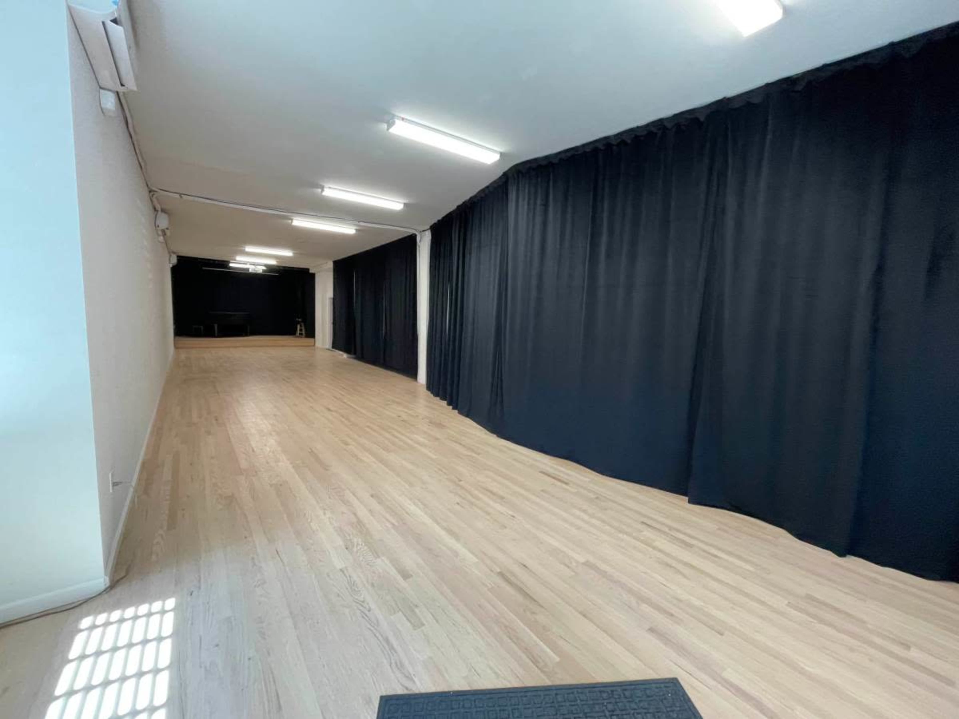 The image shows a long, narrow room with wooden flooring and black curtains lining one side.