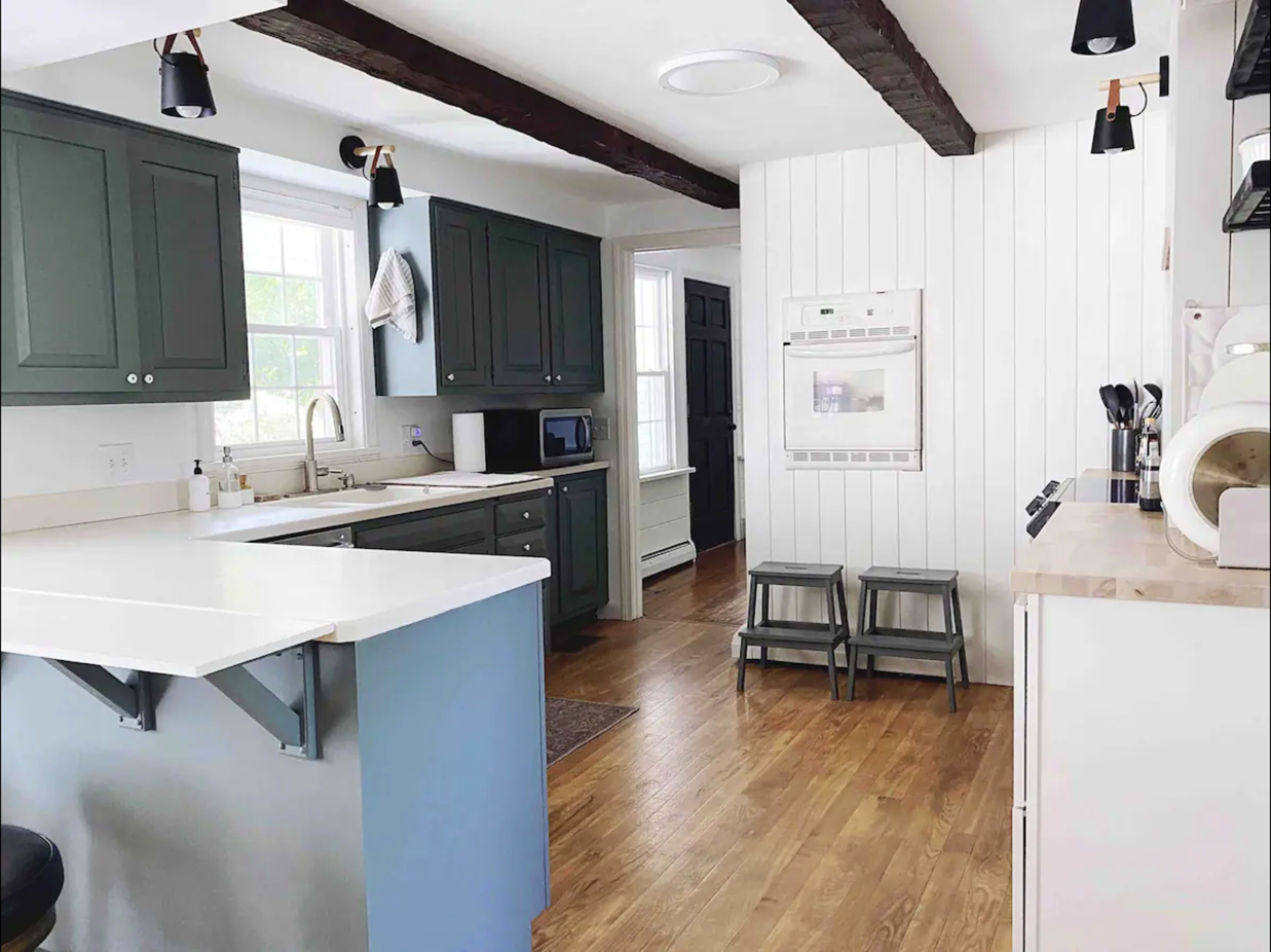 The kitchen features dark green cabinets, white countertops, and wooden flooring, with a separate area for seating and two step stools.