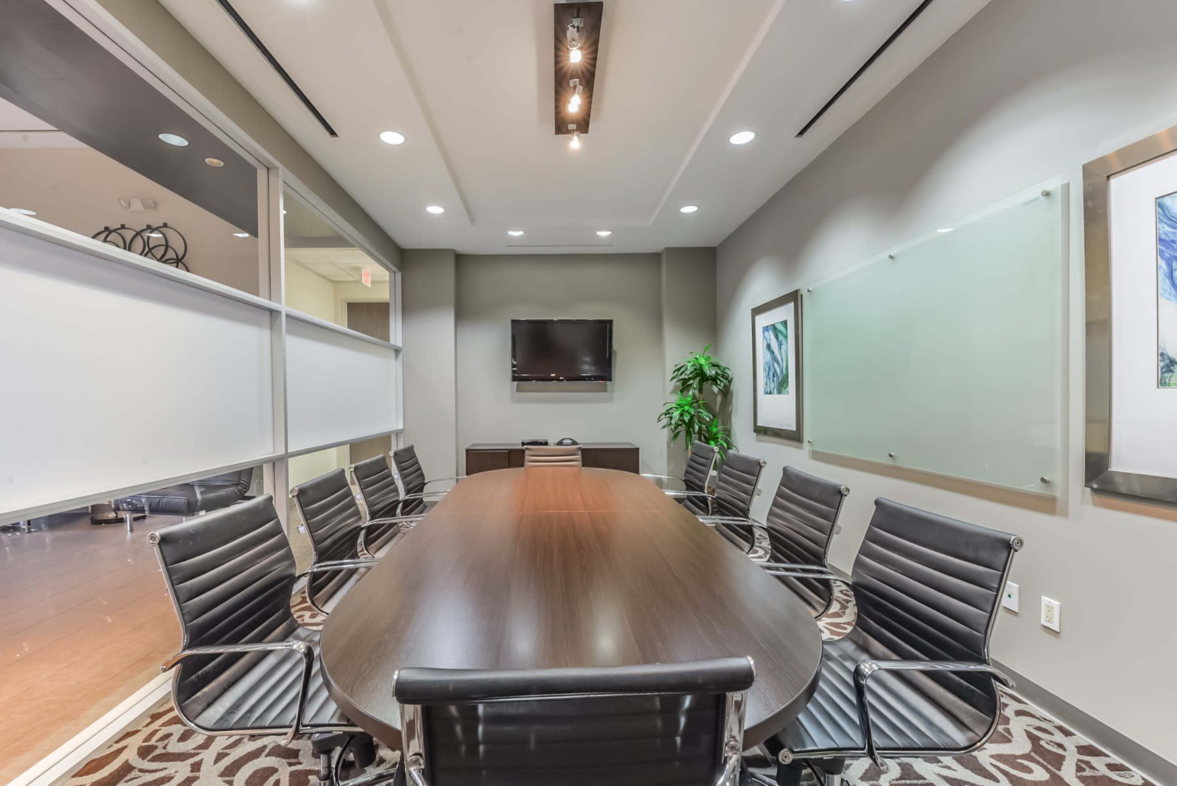 A modern conference room features a large wooden table surrounded by black leather chairs, with a television mounted on the wall and glass partitions.