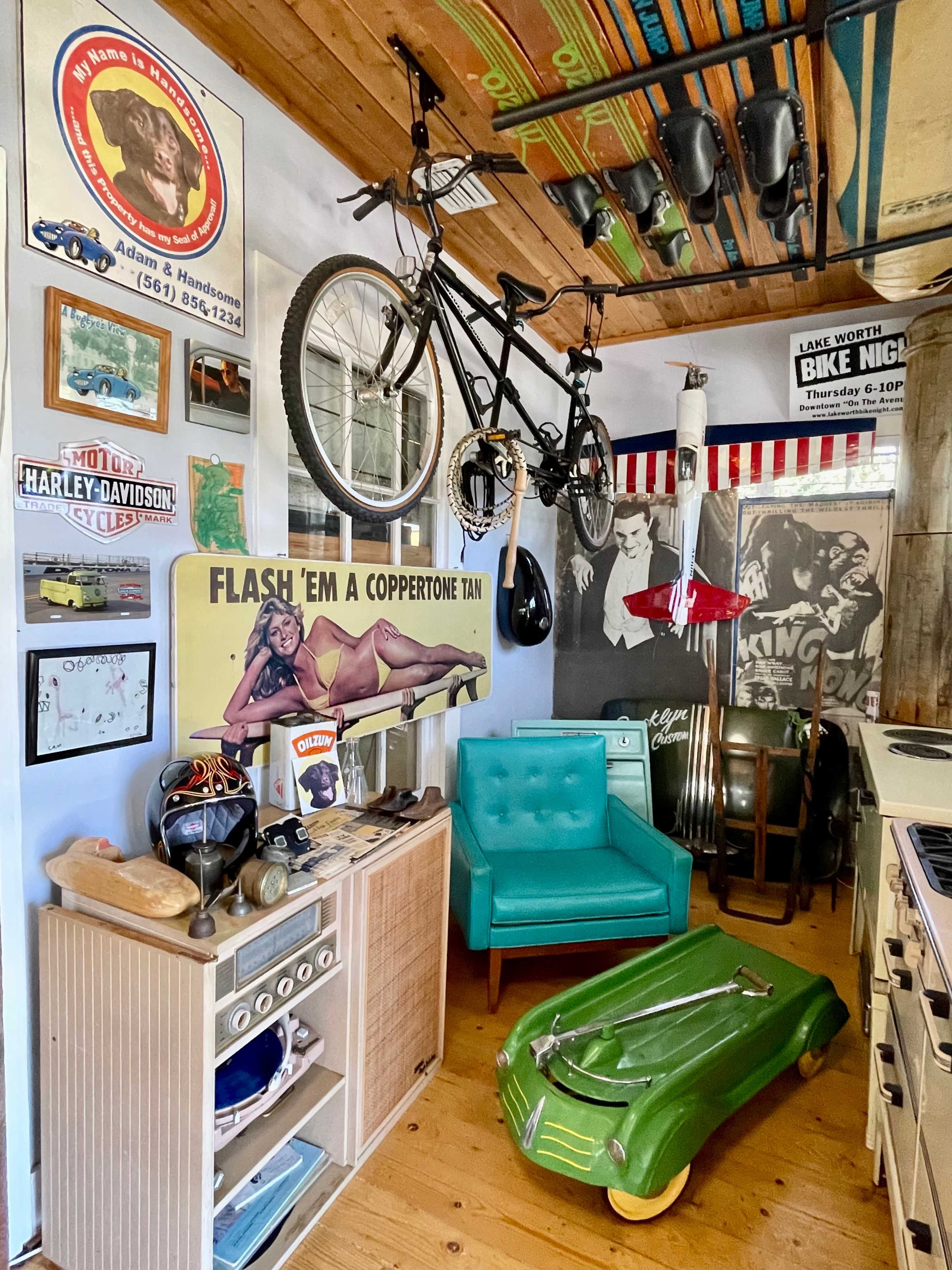 A vintage-themed room featuring a turquoise armchair, a green toy car, and various retro posters and memorabilia on the walls, along with bicycles and helmets suspended from the ceiling.