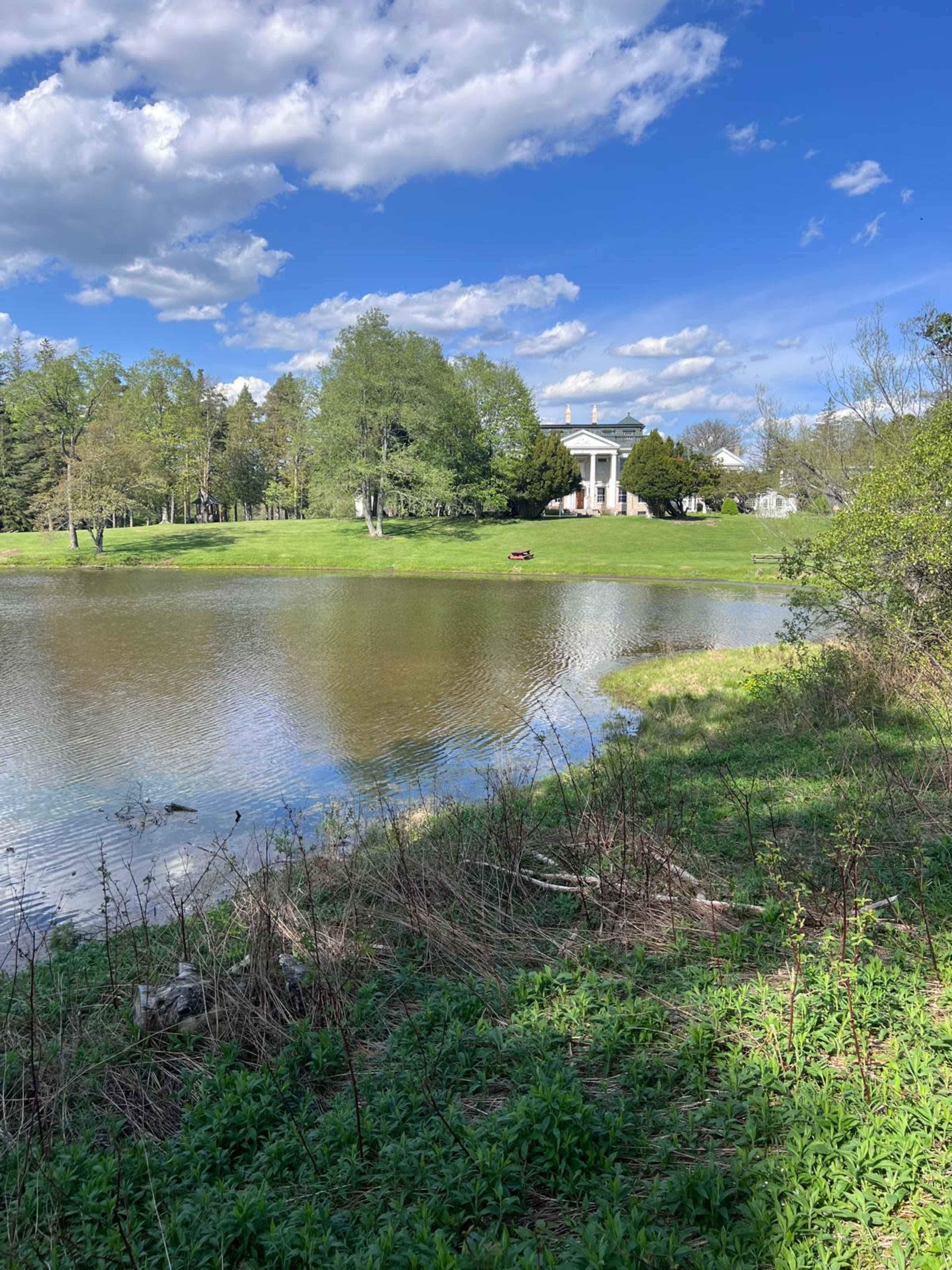 A white building with columns stands beside a pond surrounded by greenery and trees under a partly cloudy sky.