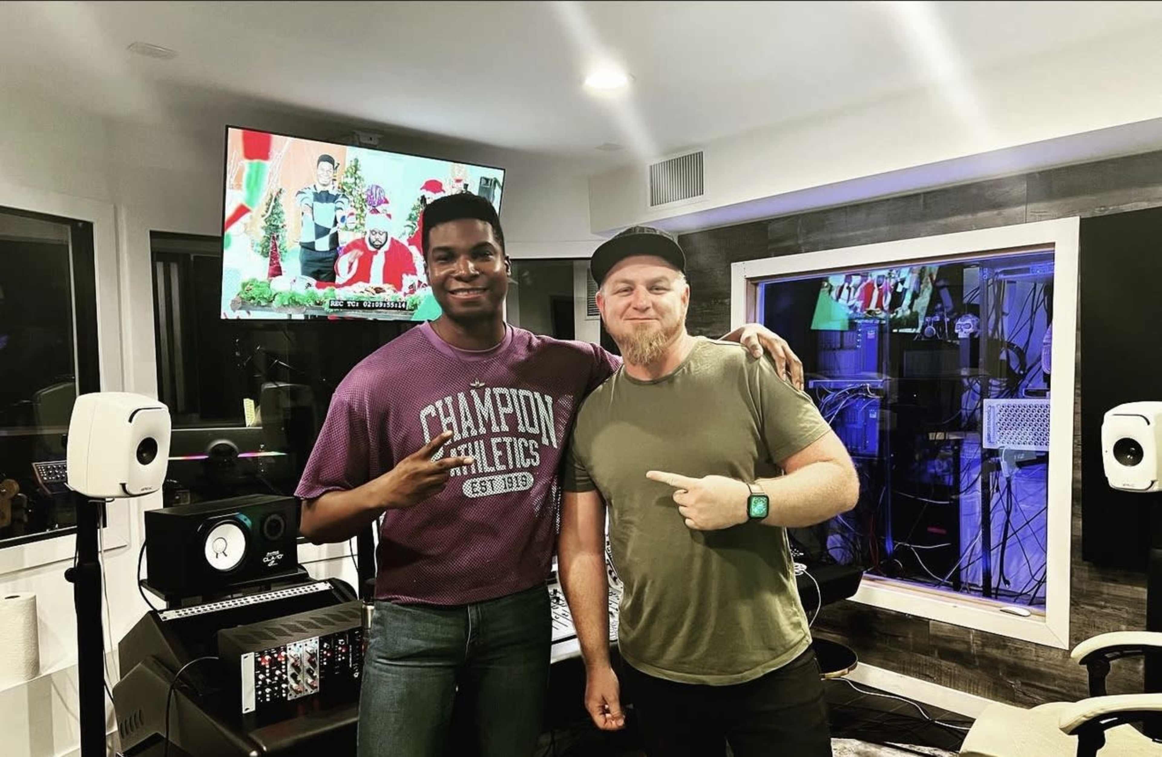 Two men pose for a photo in a well-lit recording studio, with music equipment and a television displaying a colorful scene in the background.