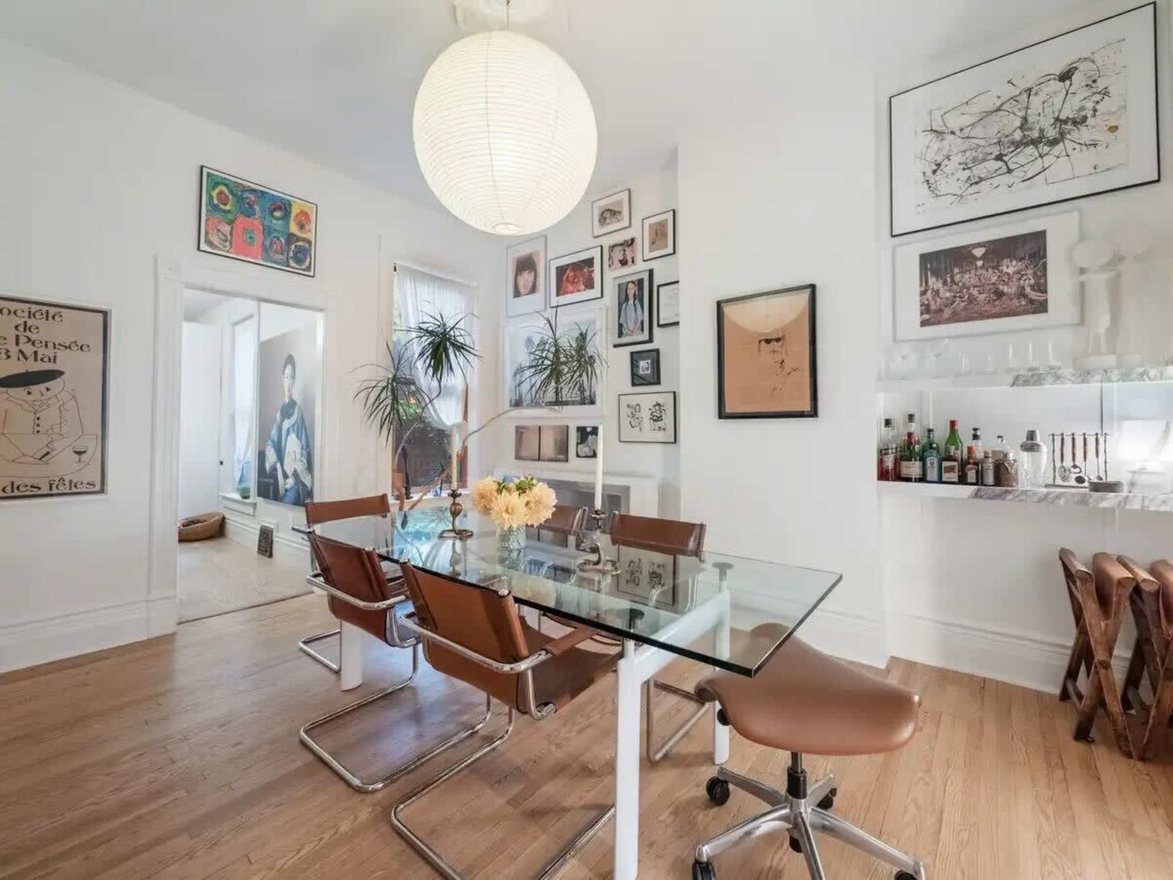 A modern dining area features a glass table surrounded by brown chairs, with walls adorned with various framed artworks and a large pendant light hanging from the ceiling.