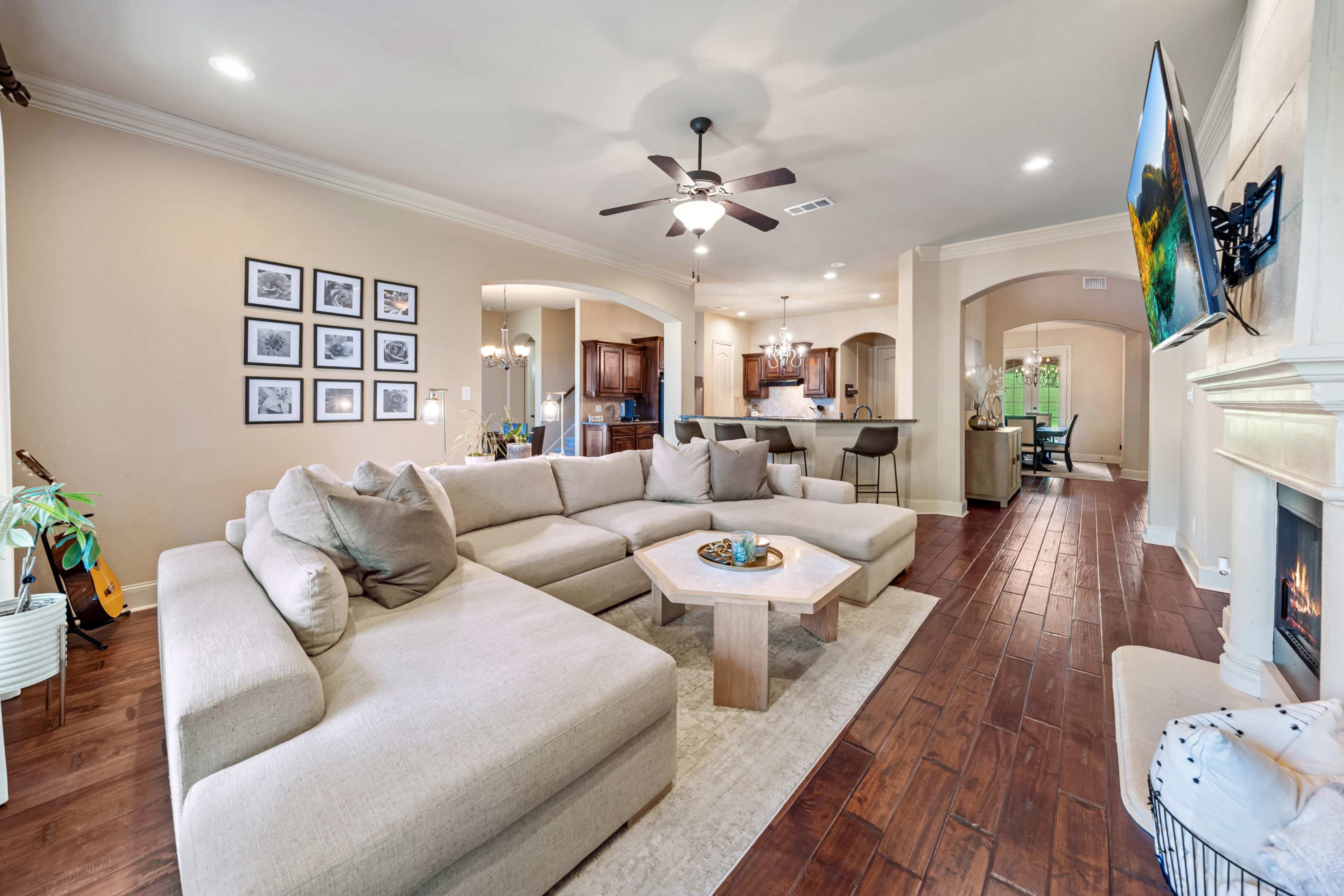 The spacious living room features a large sectional sofa, a wooden coffee table, and a television mounted on the wall, with an open layout leading to the kitchen and dining area.