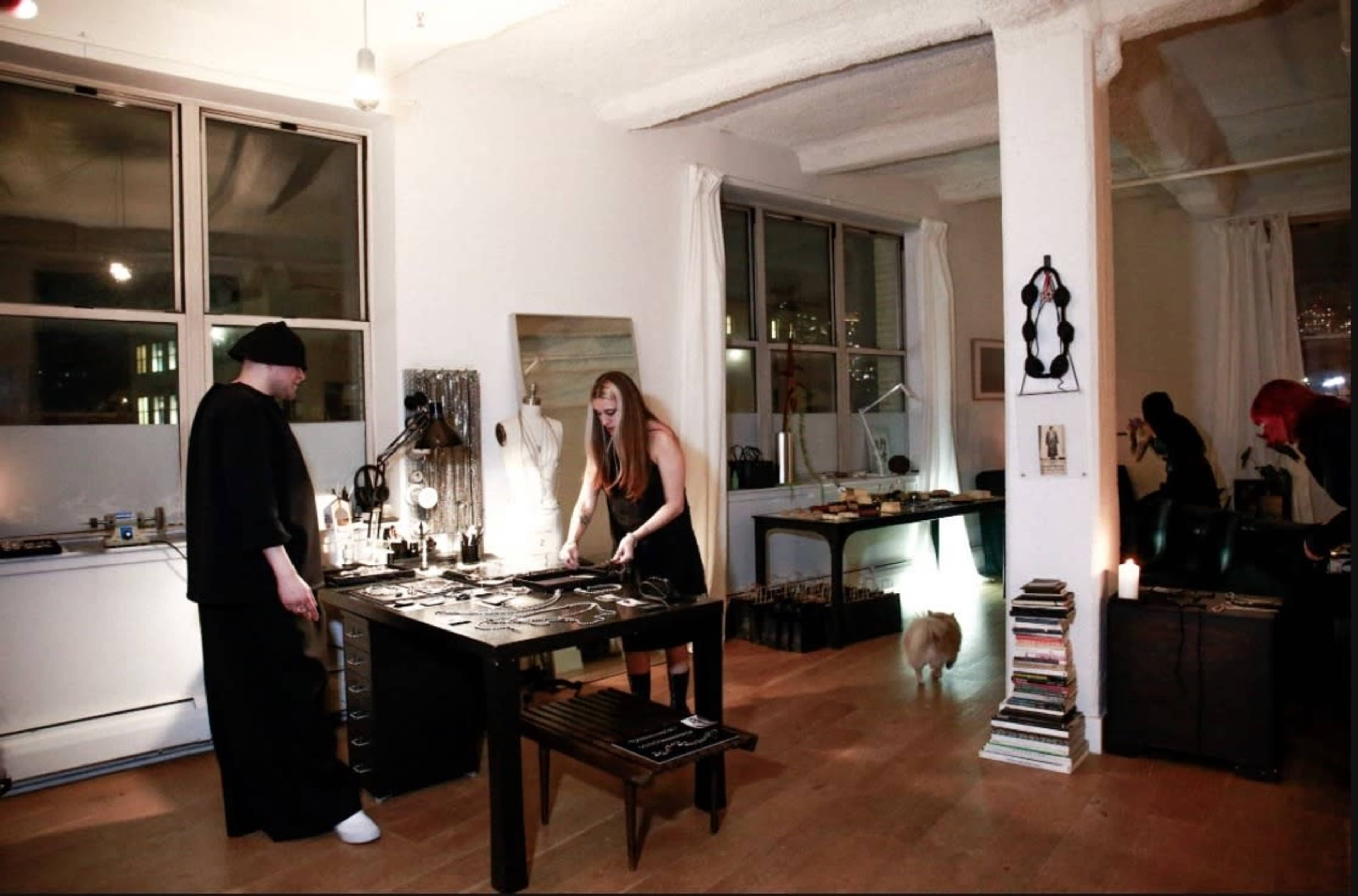 A woman arranges items on a table while a man stands nearby in a well-lit studio space with large windows.