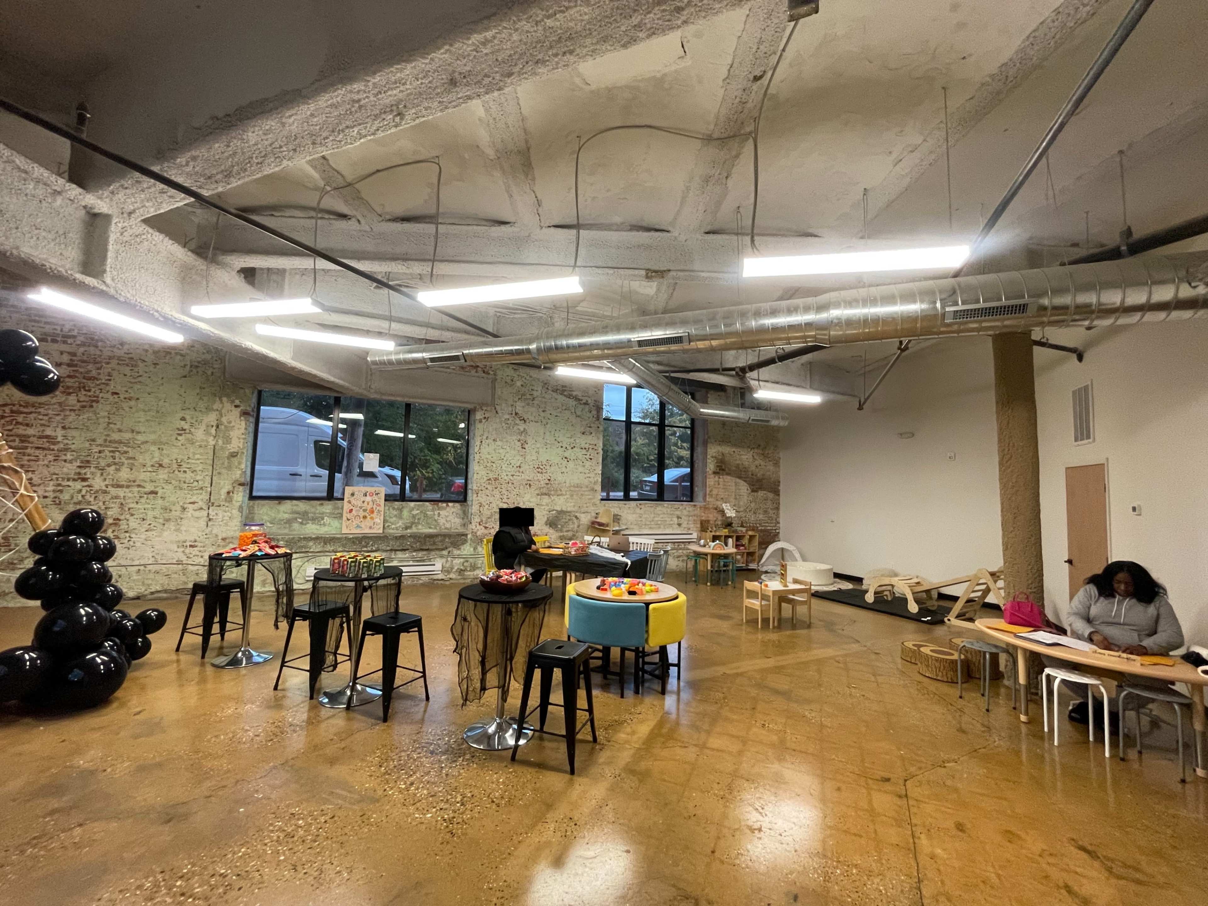 An industrial-style interior space with exposed brick walls and concrete flooring, featuring tables with fruit arrangements and a few people seated and engaged in activities.