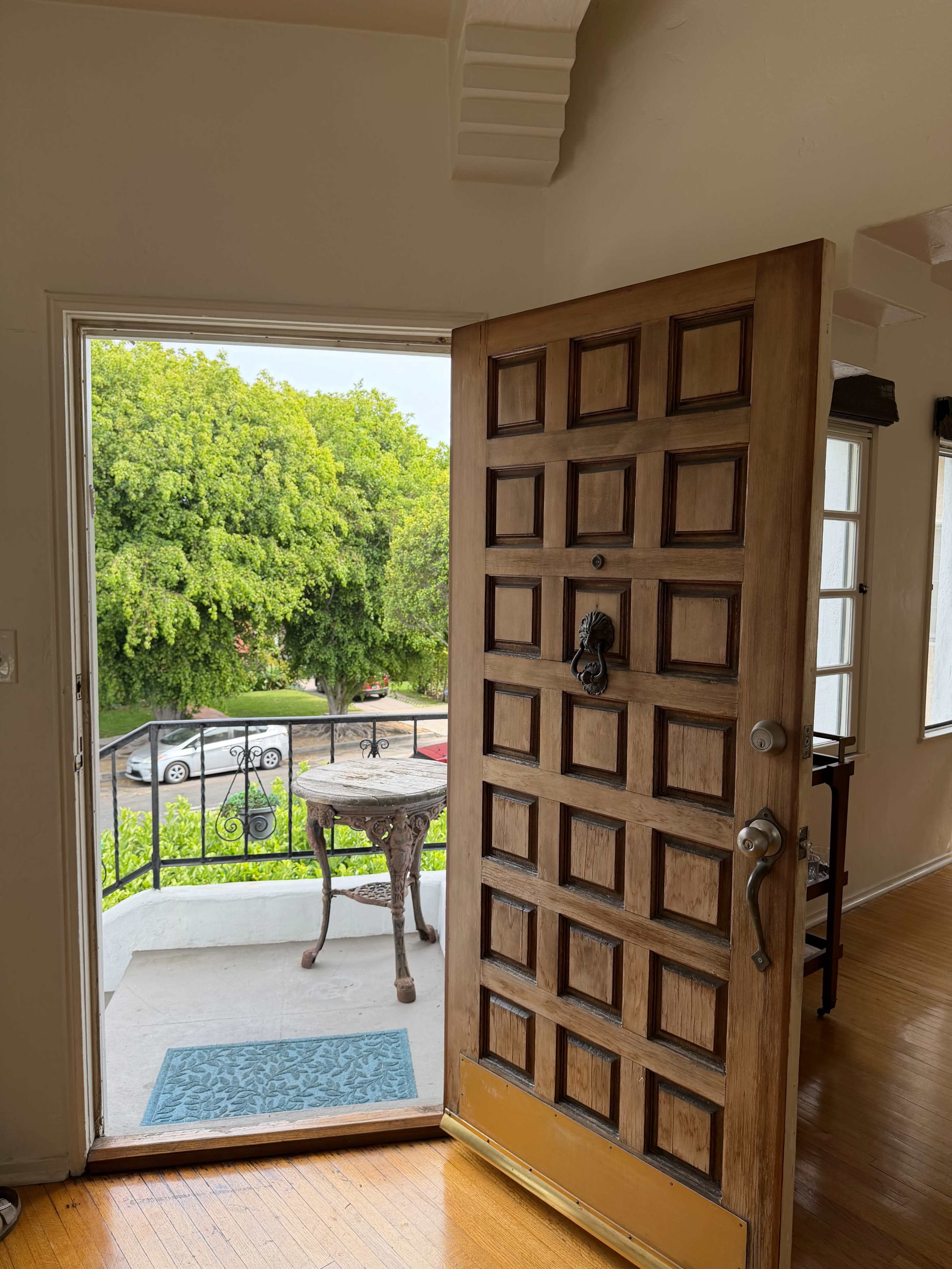 A large wooden door stands open, revealing a view of a green tree and a small table outside on a balcony.