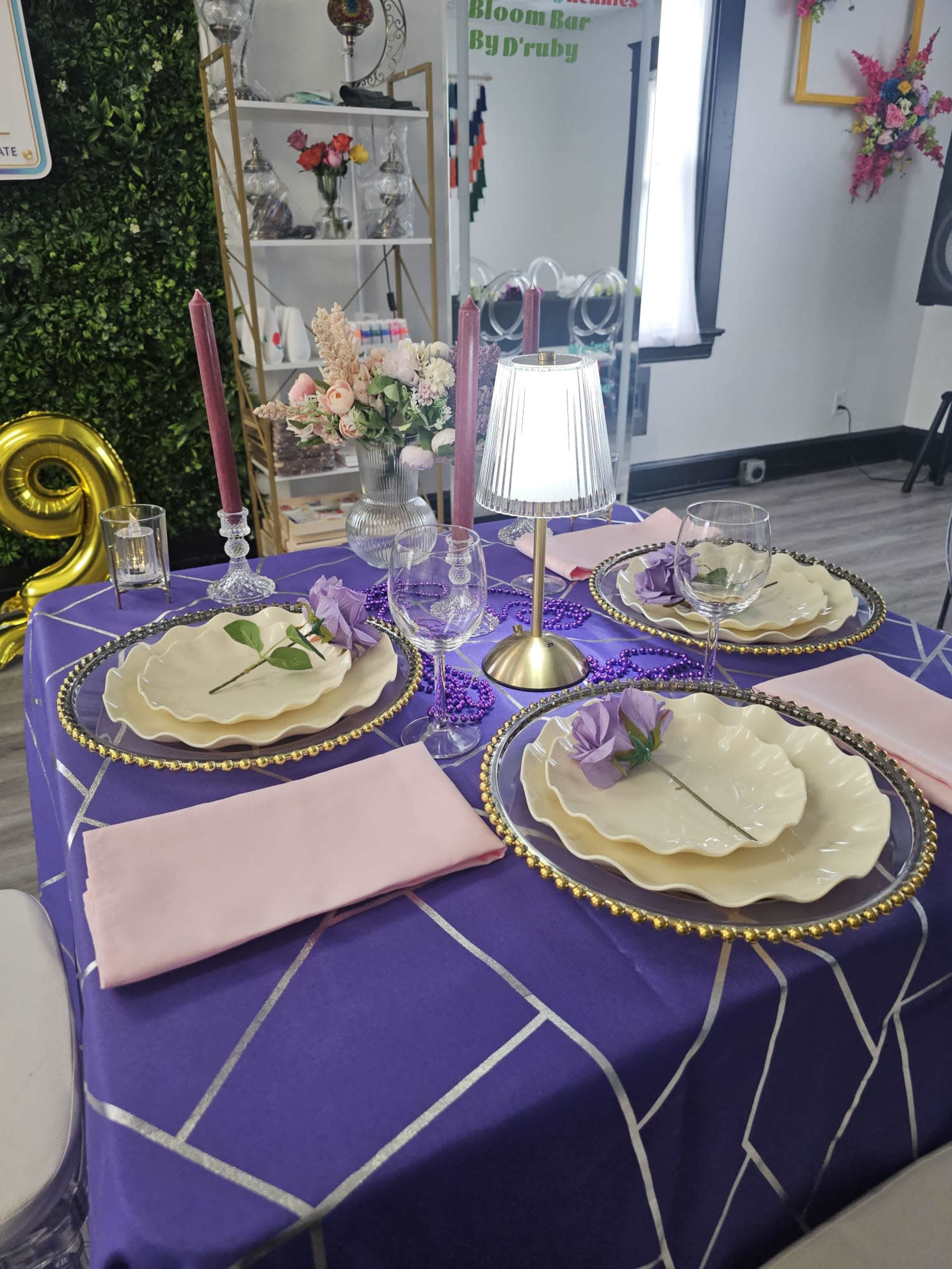 The image shows a elegantly set dining table with grey and white plates, pink napkins, glassware, and decorative candles, all arranged on a purple tablecloth.