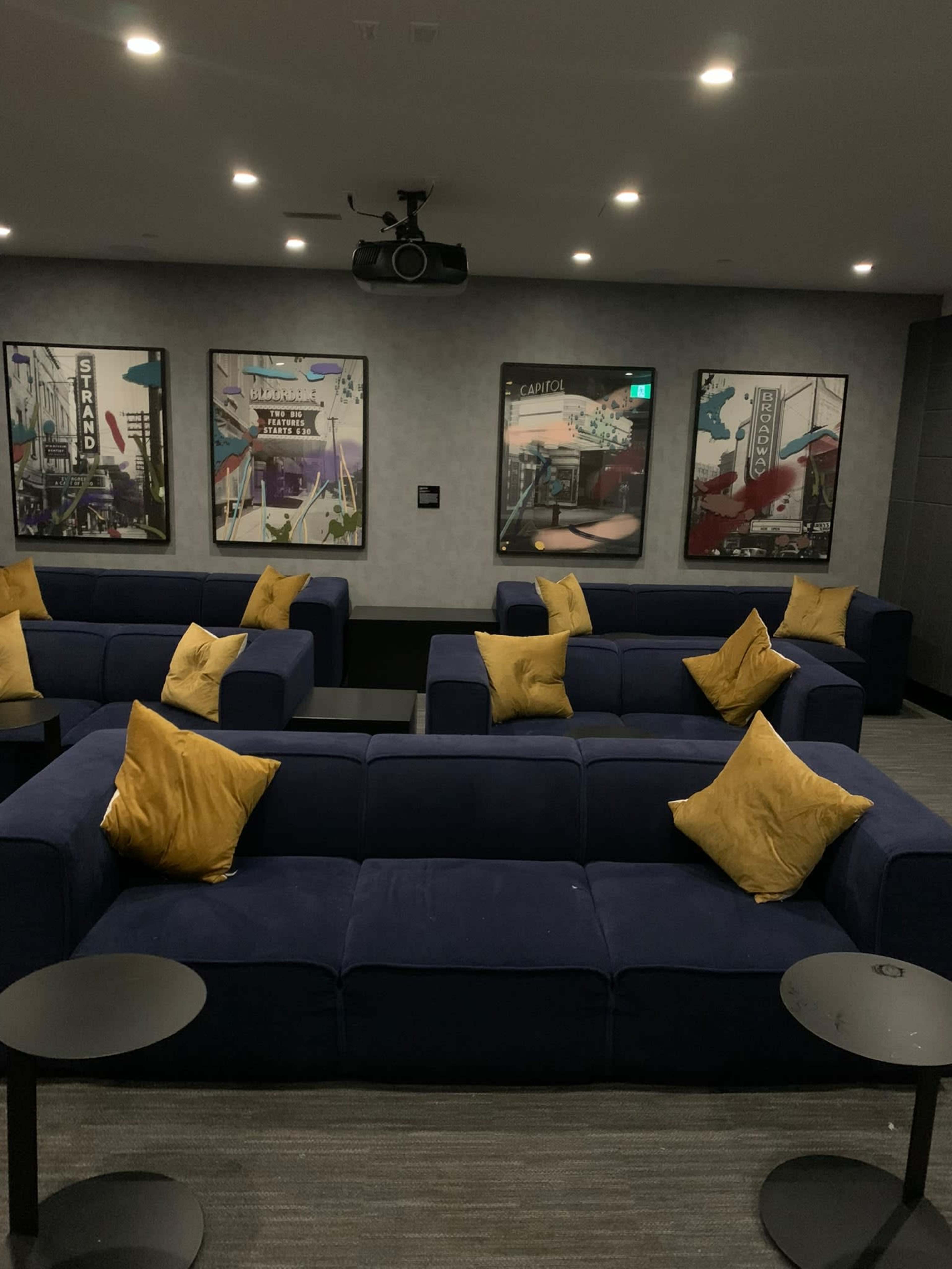 The image shows a modern lounge area with dark blue sofas adorned with yellow pillows, surrounded by framed artwork on the walls and small black tables.