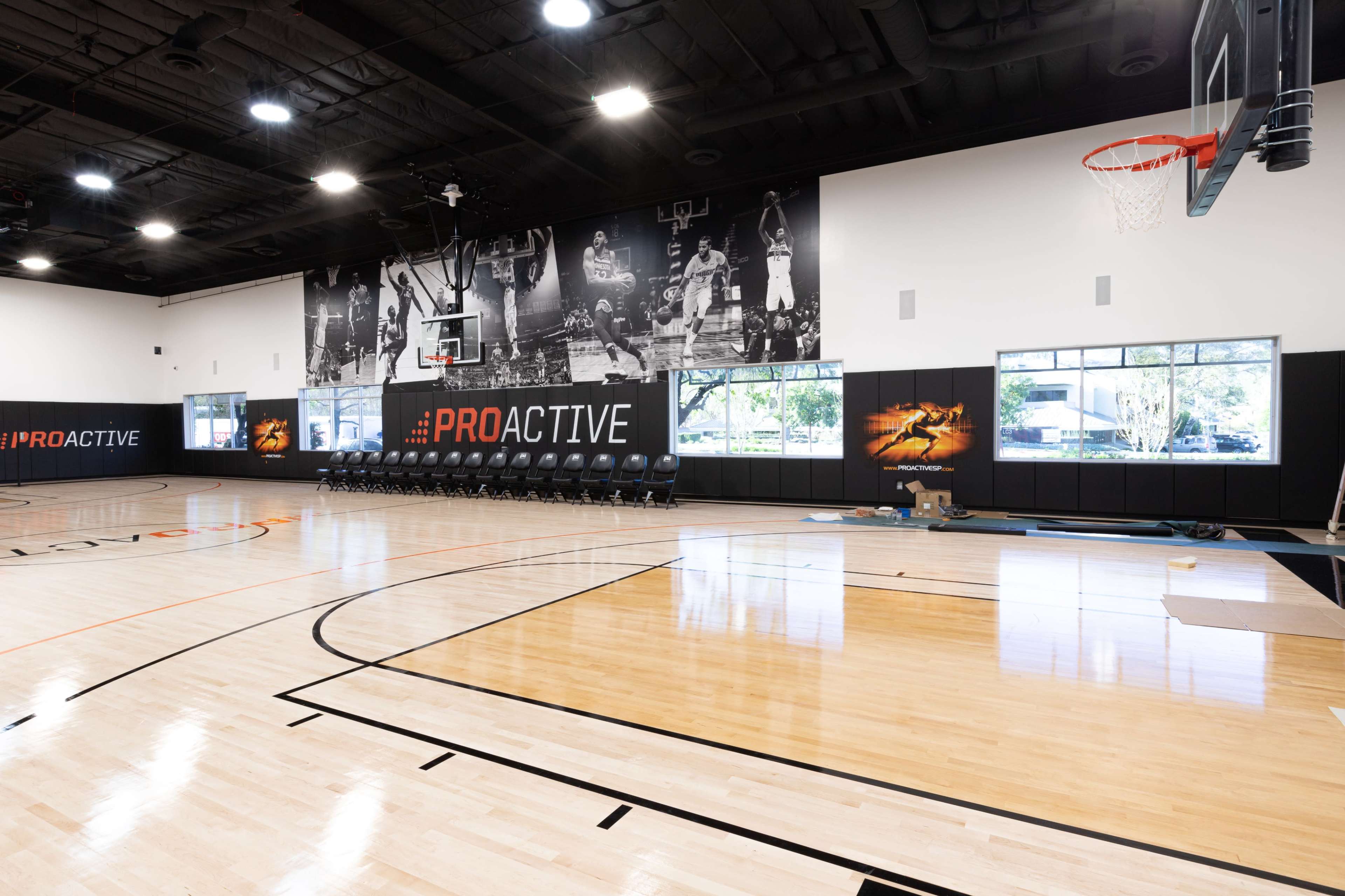 The image shows a basketball gymnasium featuring a polished wooden floor, a hoop, and a large mural of basketball players on one wall.