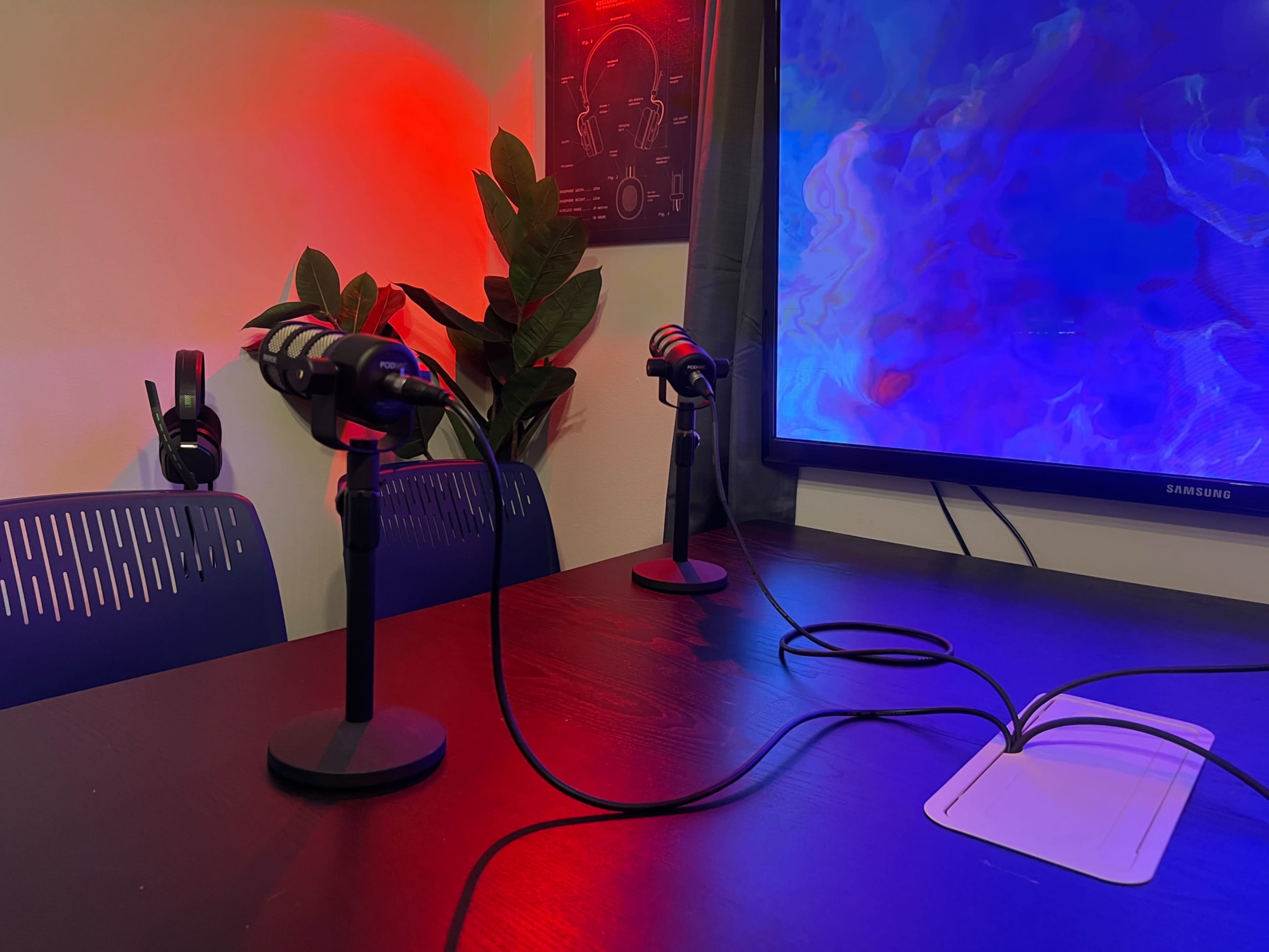 Two microphones on stands are positioned on a dark table in front of a large screen, with headphones on the table and red lighting creating a contrasting atmosphere.