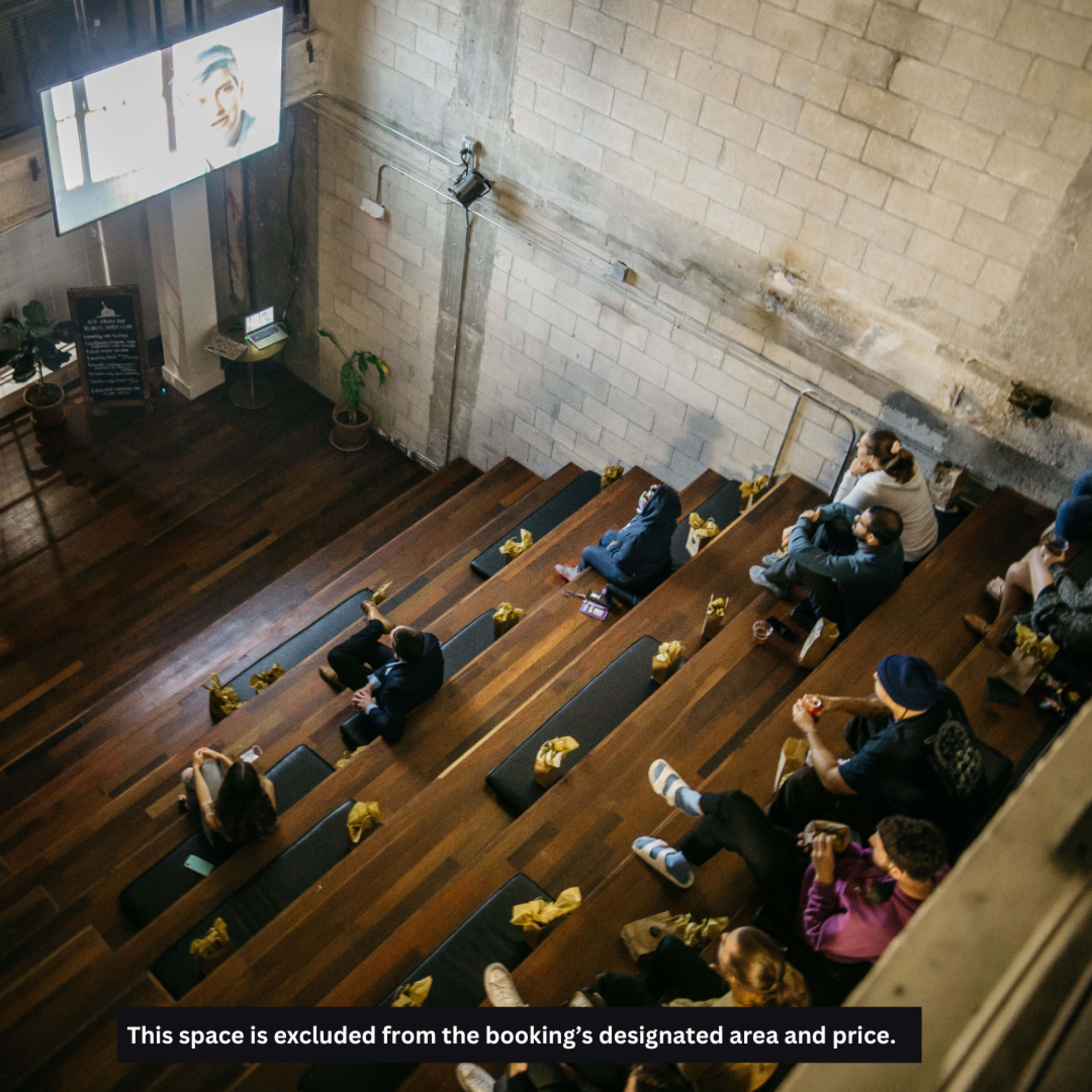 A group of people sits on wooden bleachers in a dimly lit space, watching a screen at the front of the room.
