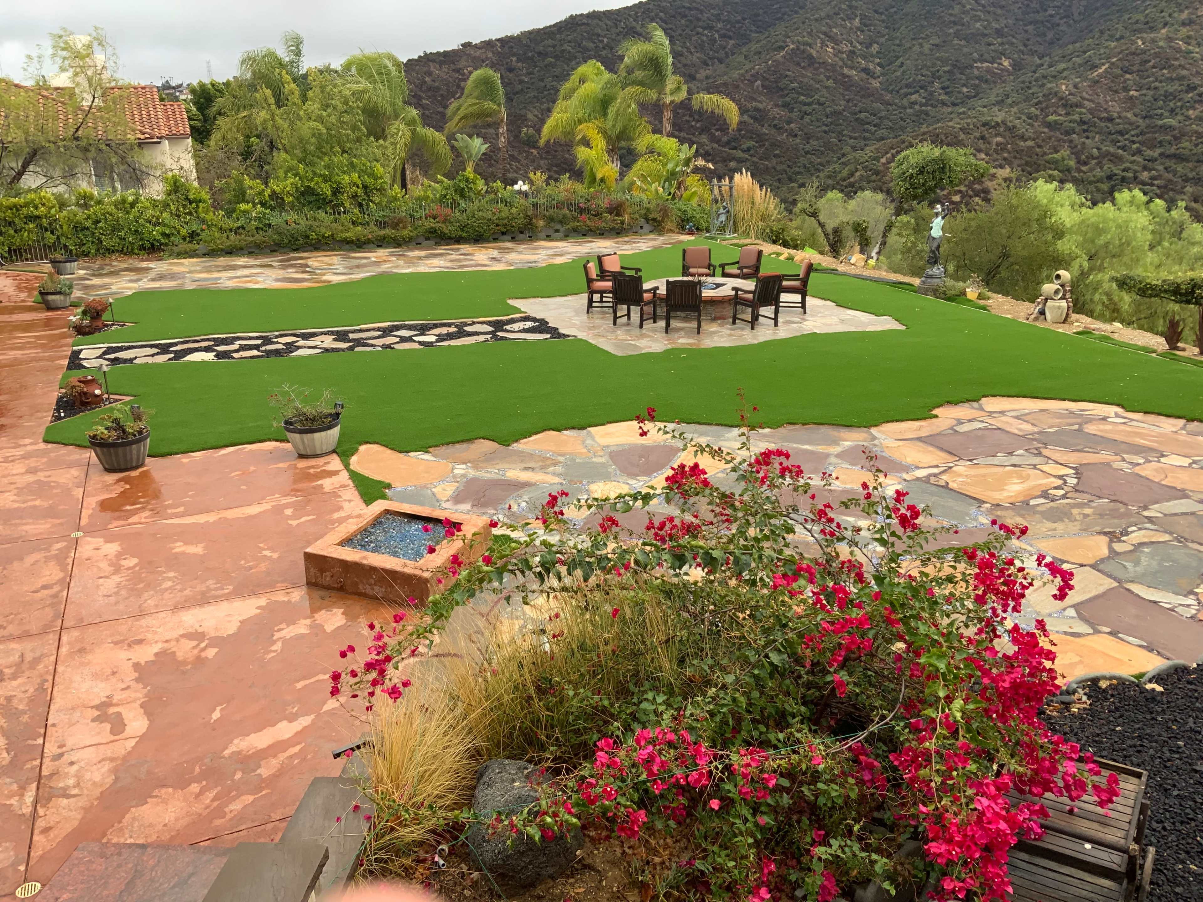 A landscaped outdoor area featuring a stone patio, a green lawn, a dining set, and blooming bougainvillea in the foreground, with hills in the background.