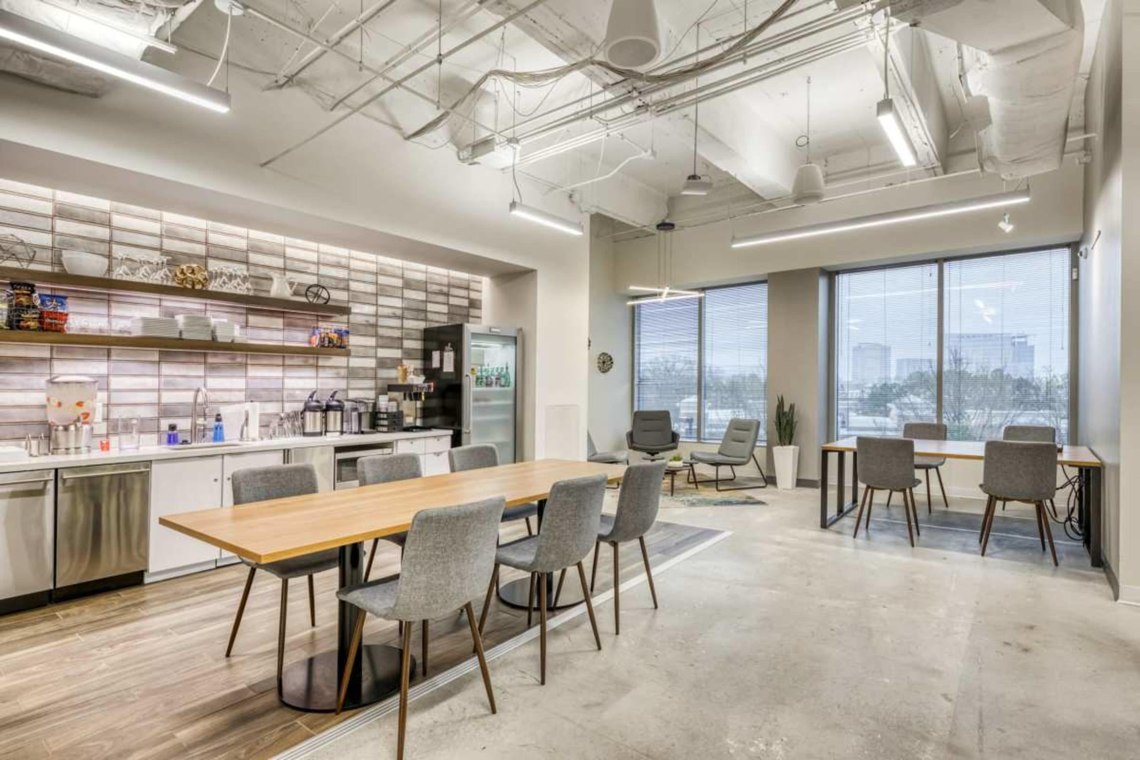 The image shows a modern office kitchen area with a long wooden dining table, gray chairs, and large windows overlooking a cityscape.