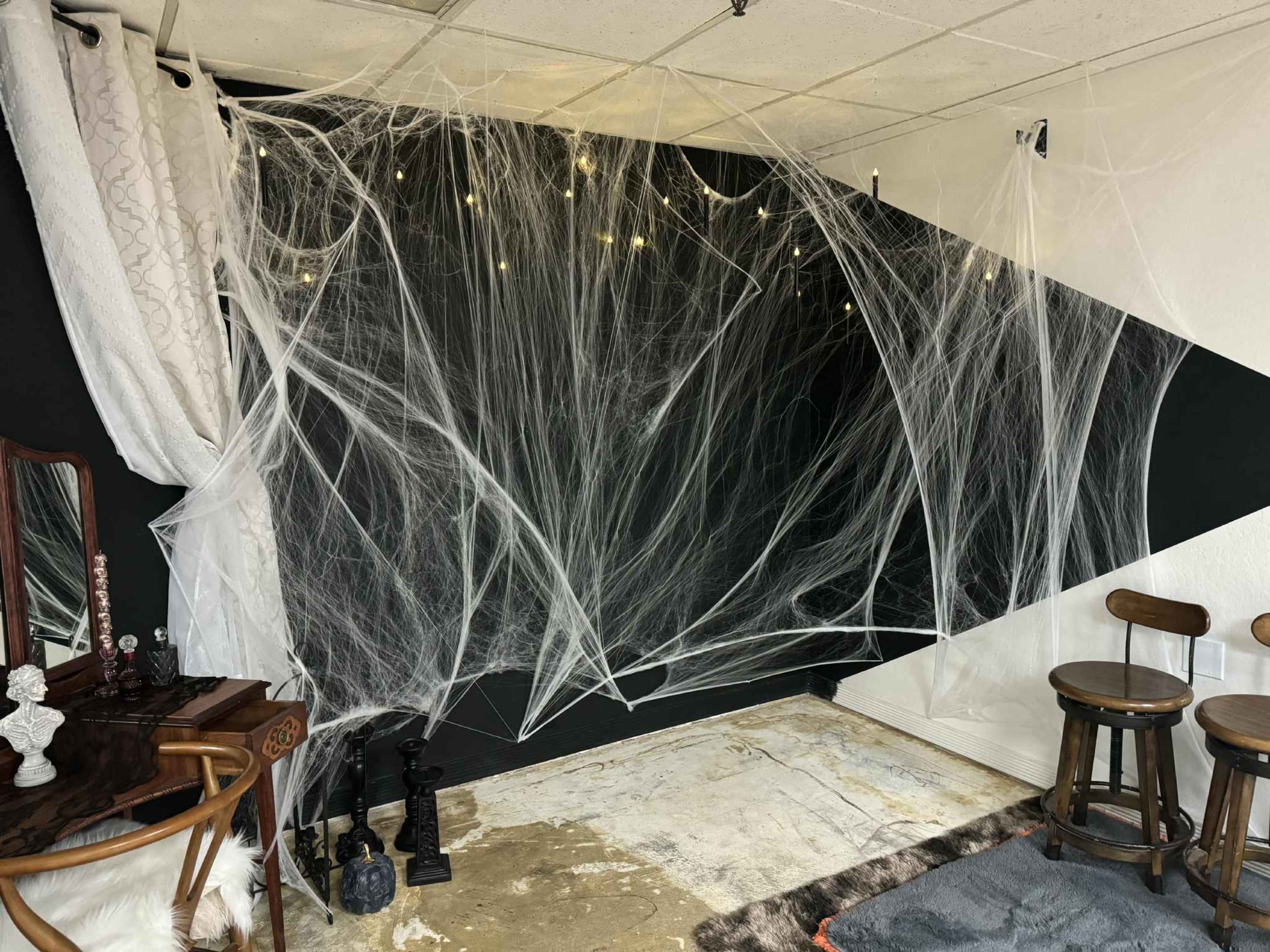 The image shows a corner of a room decorated with white spider web-like material and string lights against a black wall, accompanied by a vintage-style mirror and two wooden stools.
