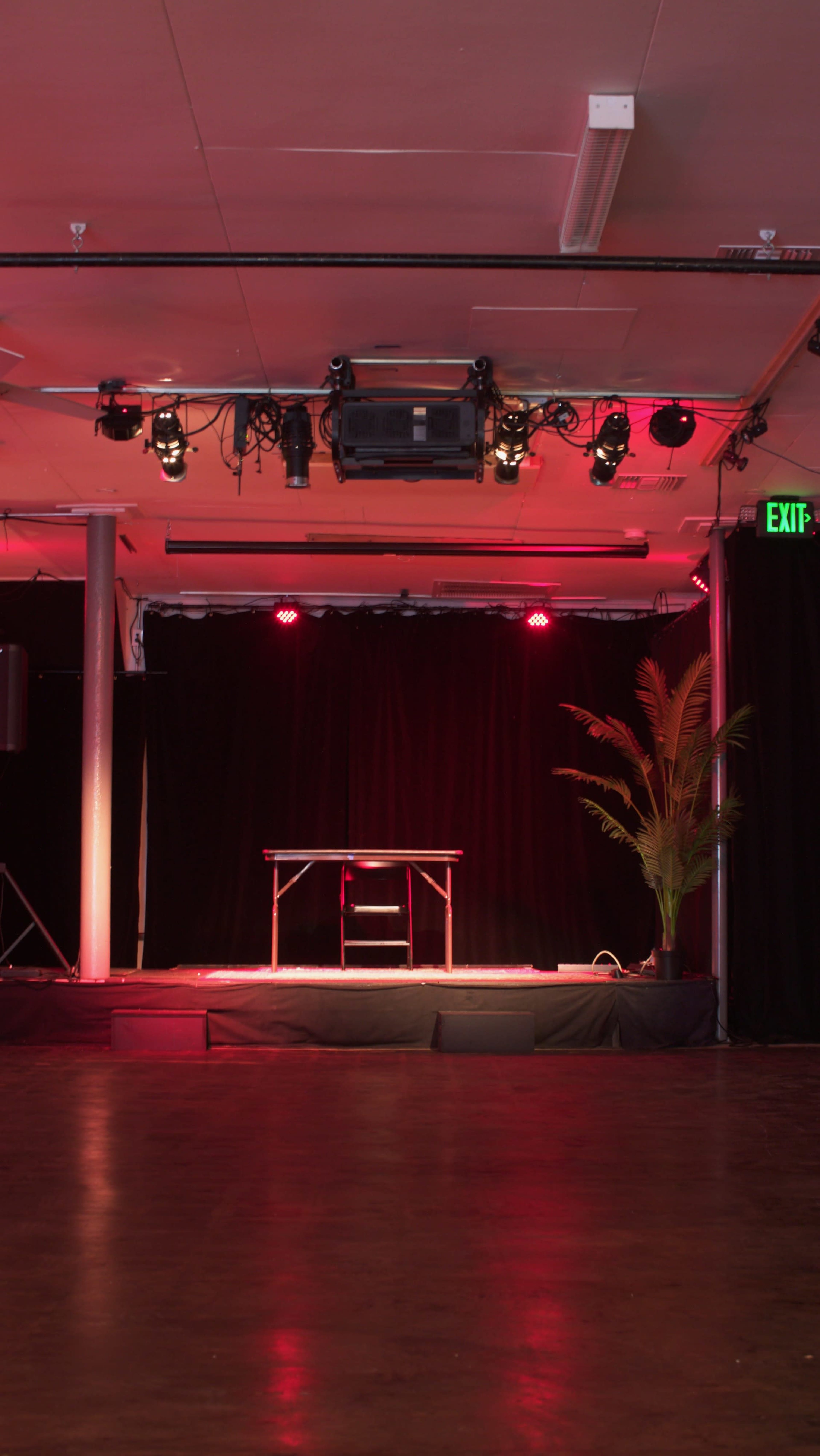 The image shows a small stage area with a black curtain backdrop, illuminated by red lights, featuring a table and a chair, and a potted plant in the corner.