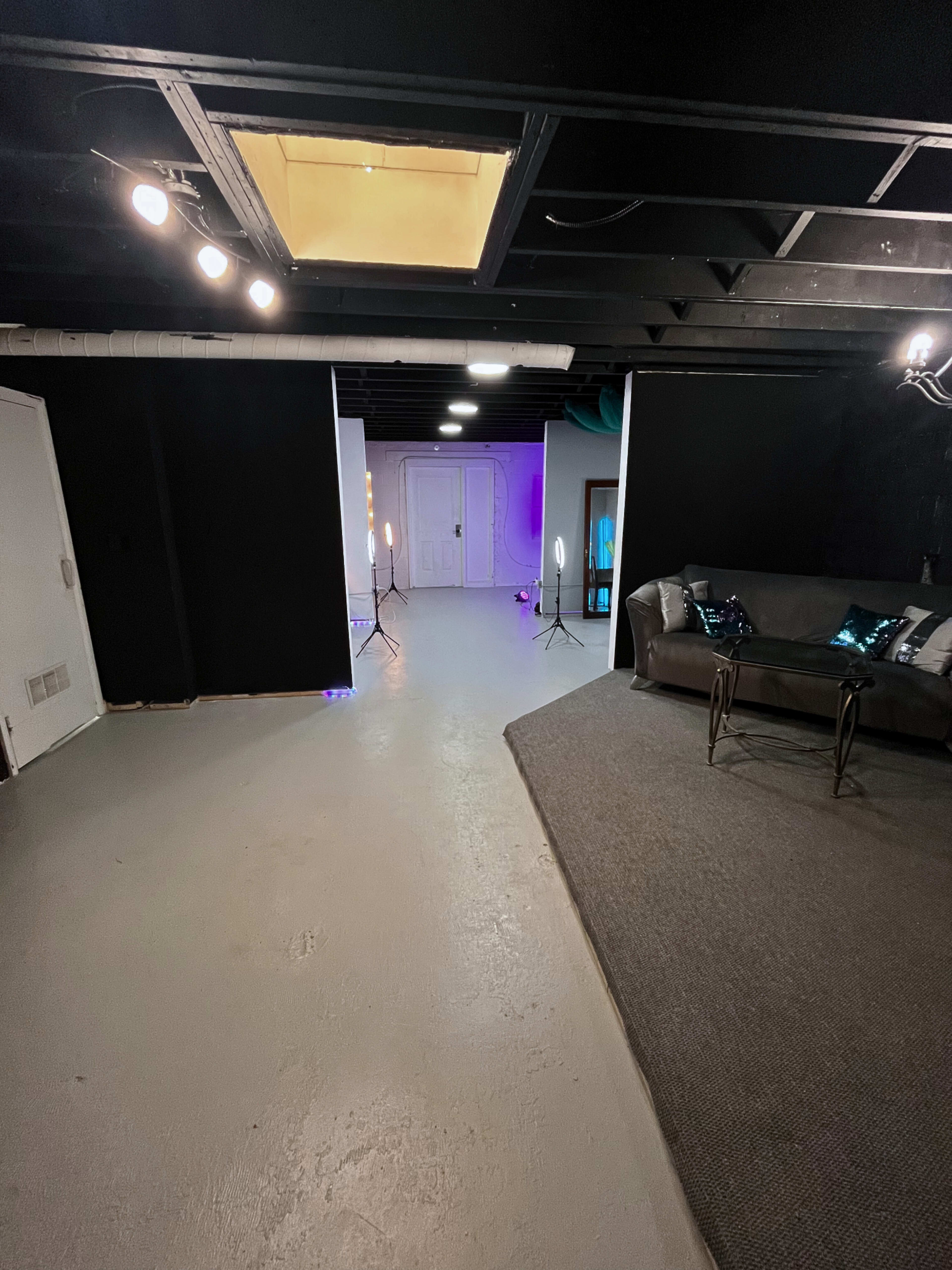 A basement room with a light-colored floor, black walls, and a couch, with lighting equipment set up in the center.