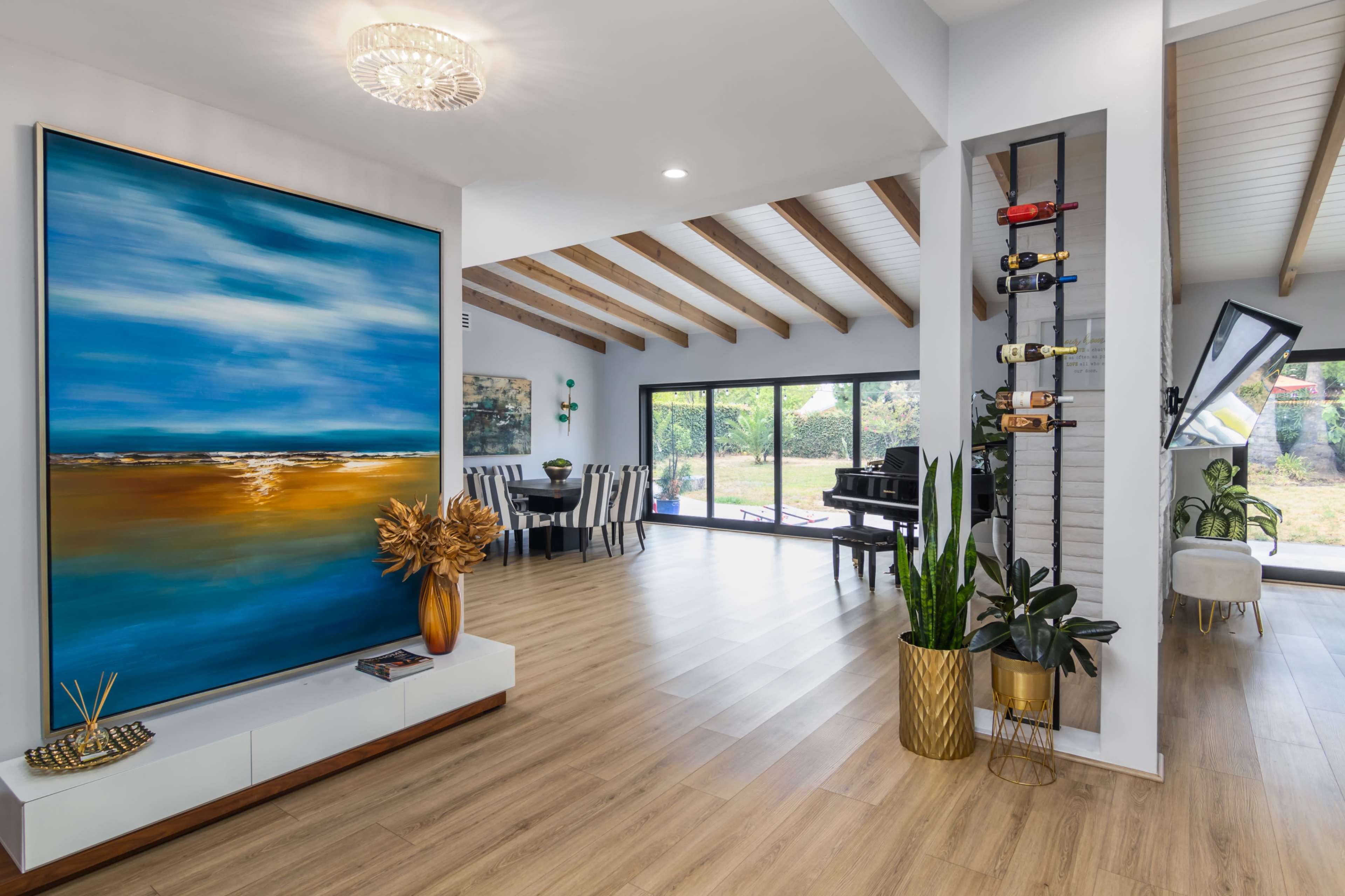 A modern living space featuring a large abstract painting, wooden beams on the ceiling, and large glass doors opening to a garden area.
