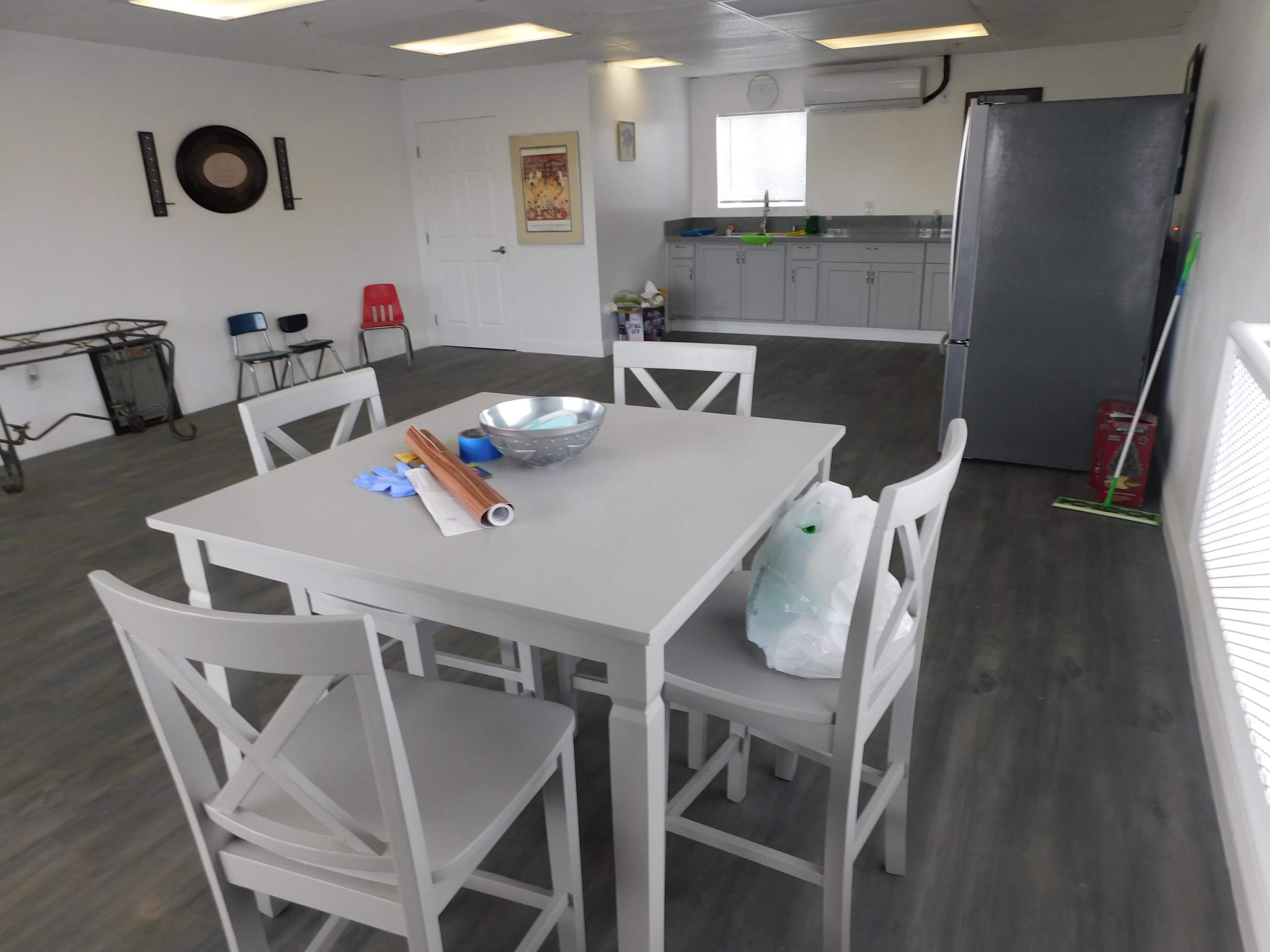 A minimalist room features a gray dining table with four chairs, a bowl on the table, and a kitchenette area in the background.