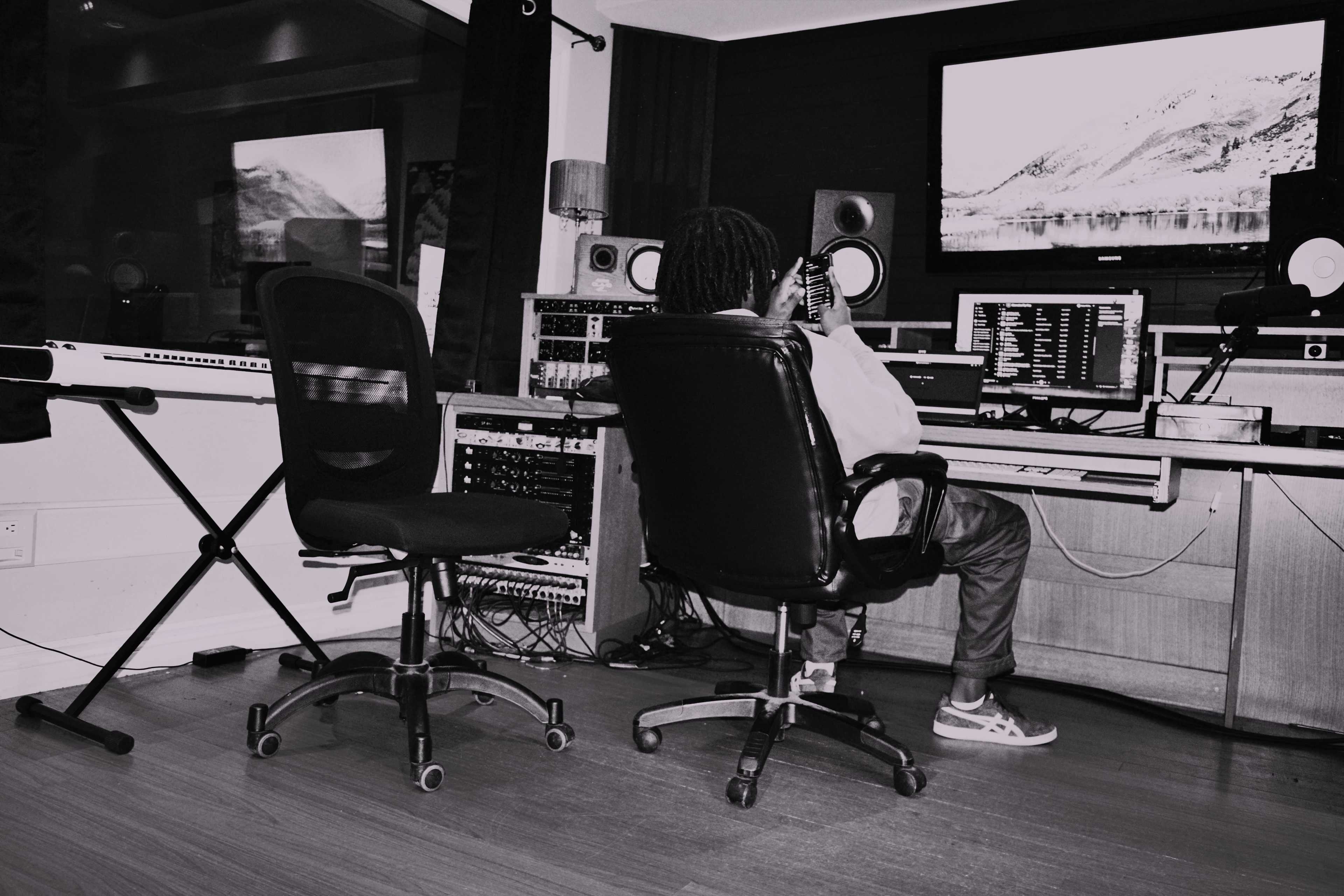 A person sits at a desk in a music studio, working with audio equipment and a computer screen displaying a landscape.