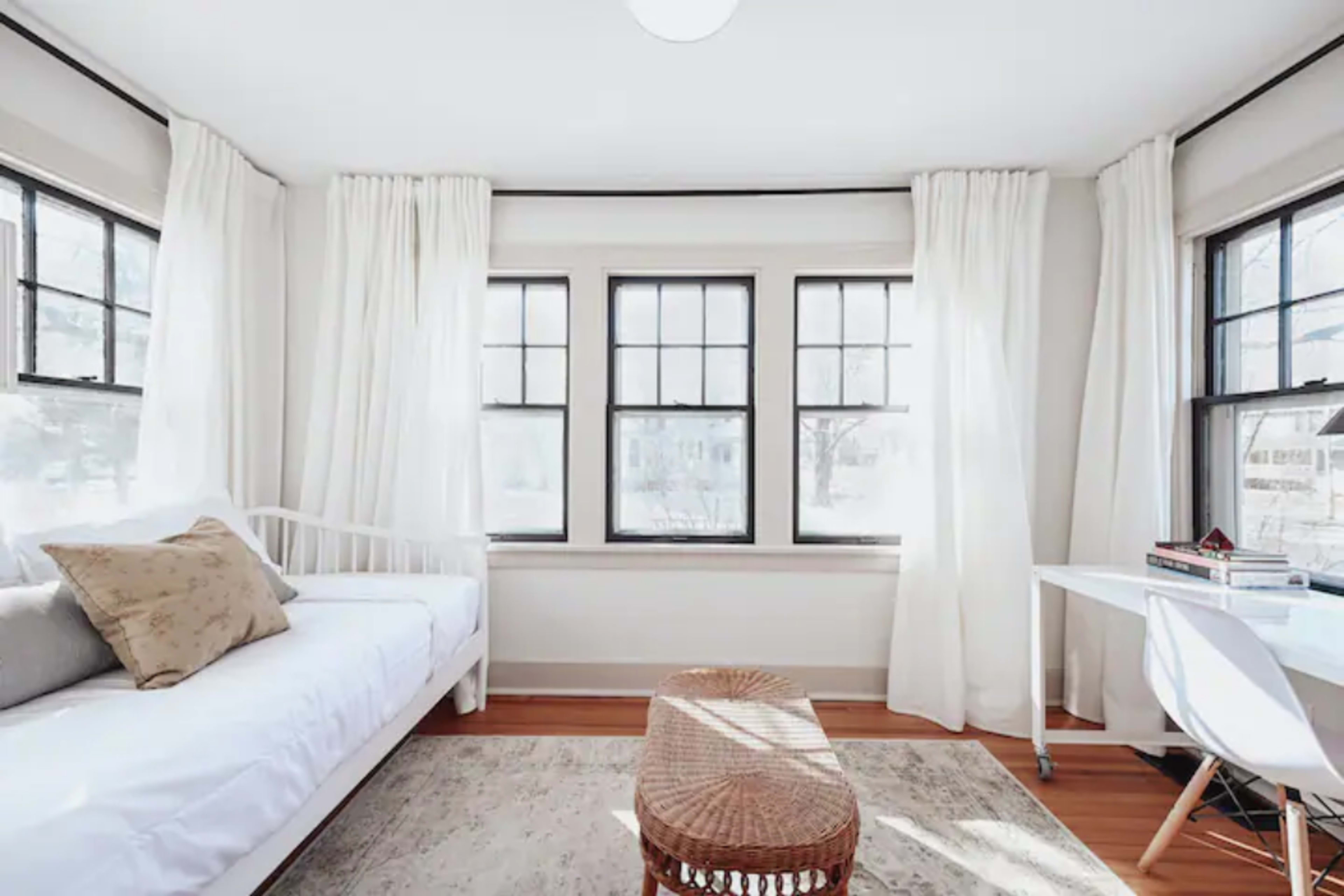 A bright, minimalist room features a daybed, a woven coffee table, and a desk, with large windows draped in white curtains letting in natural light.