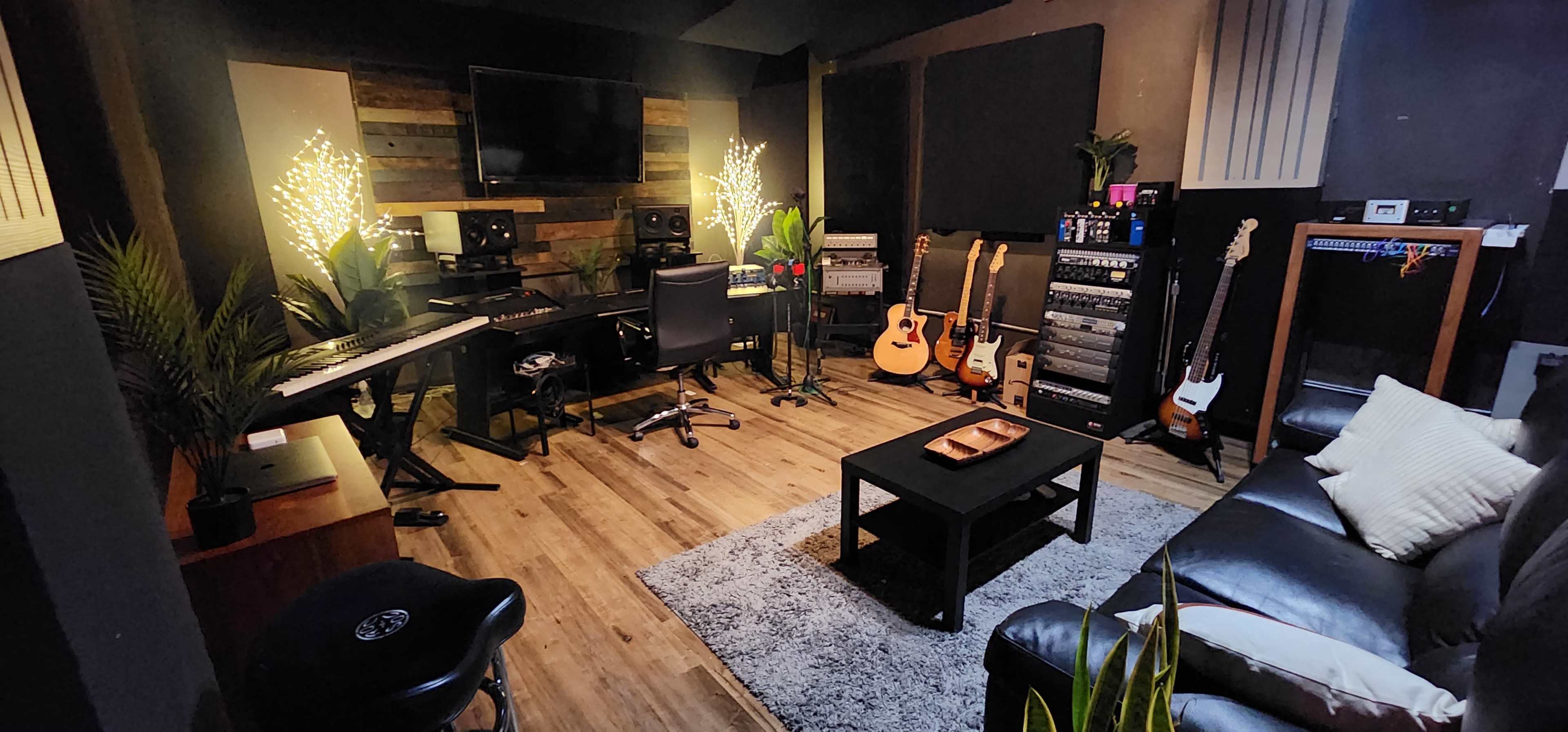 The image shows a music studio featuring a keyboard, mixing equipment, guitars, and a cozy seating area with decorative lights and plants.