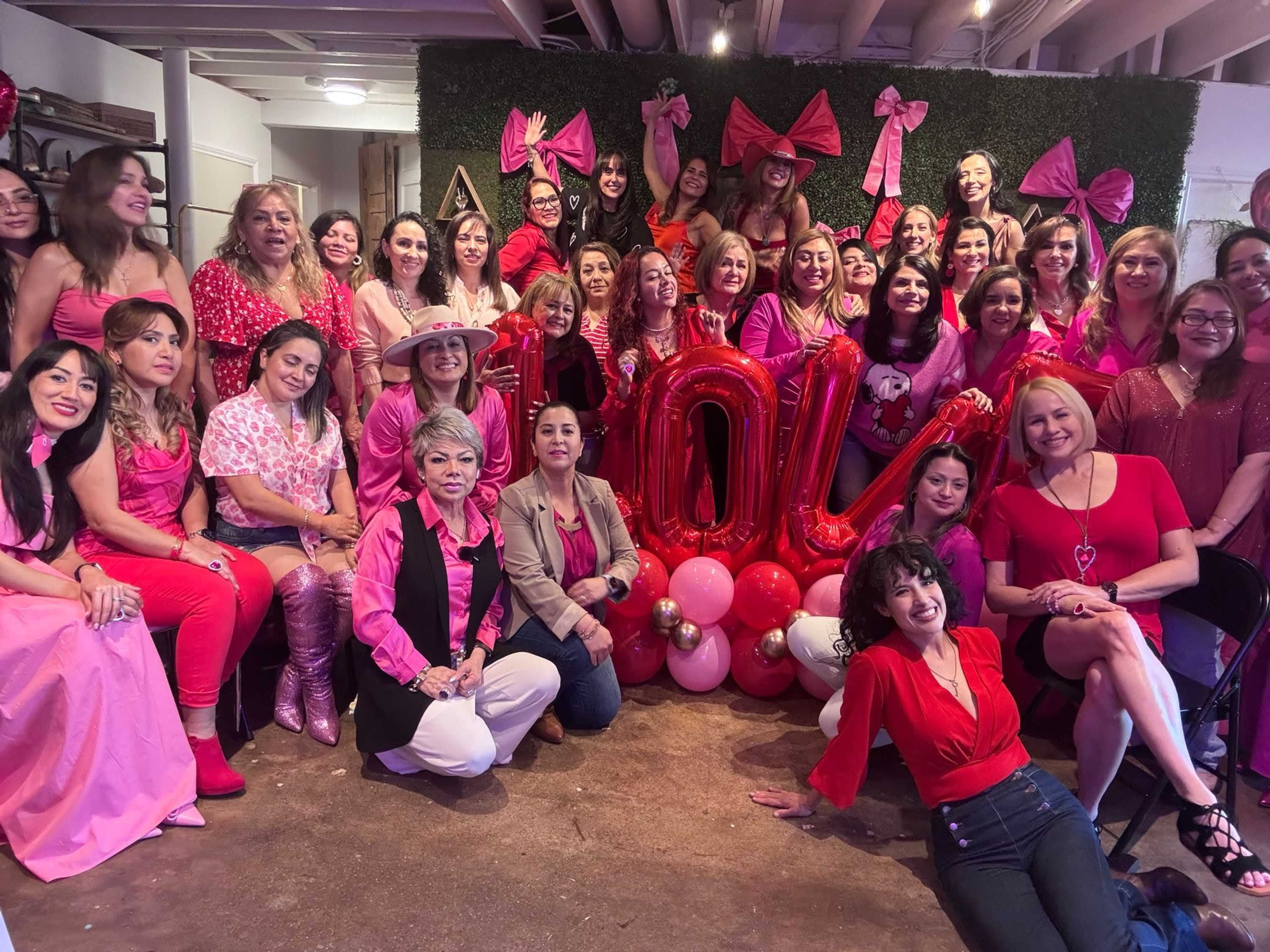 A group of women dressed in red and pink attire poses for a photo in a decorated indoor space.