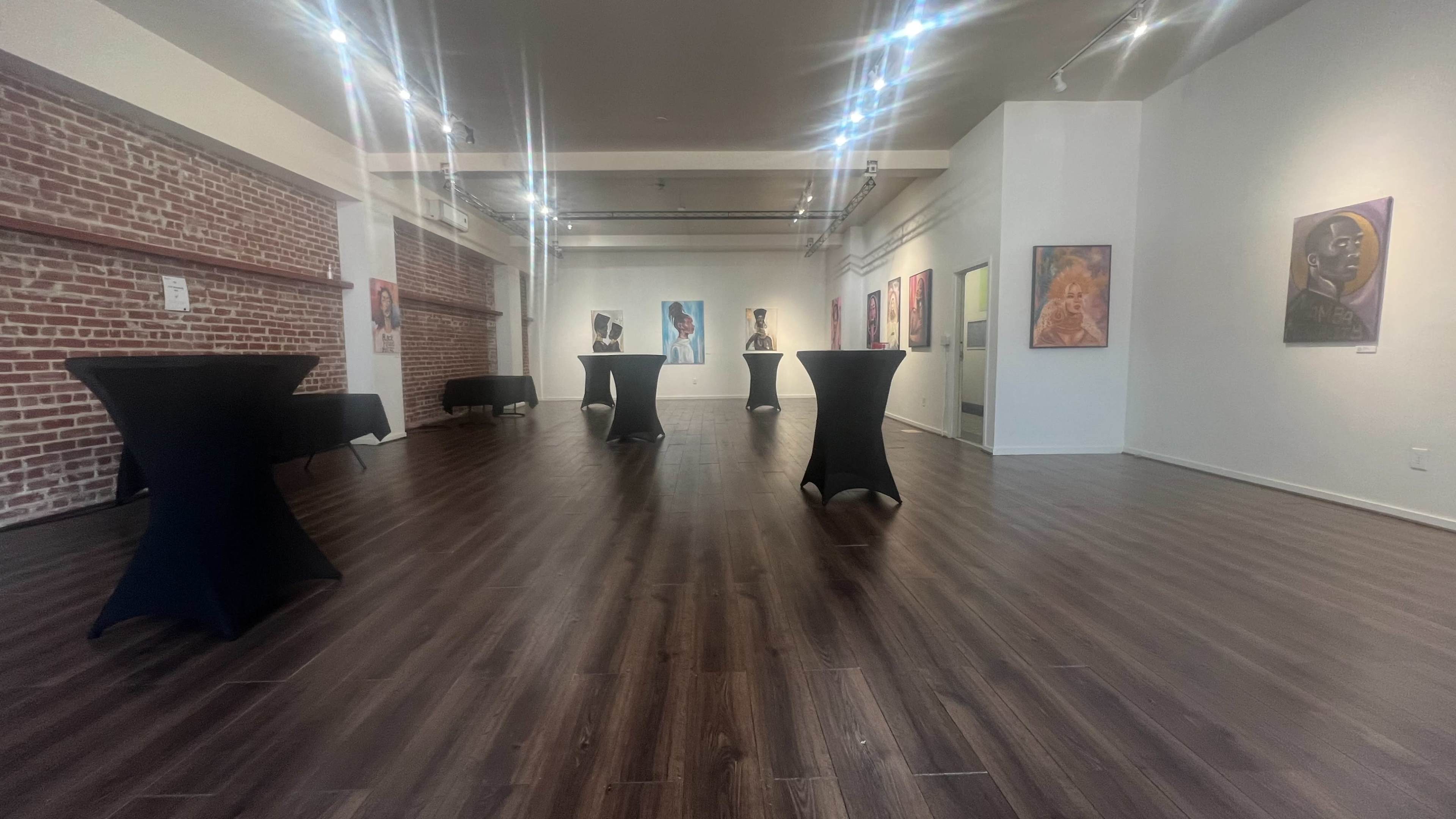 An art gallery with black cocktail tables set up on a polished wooden floor, surrounded by various paintings hanging on the white walls.