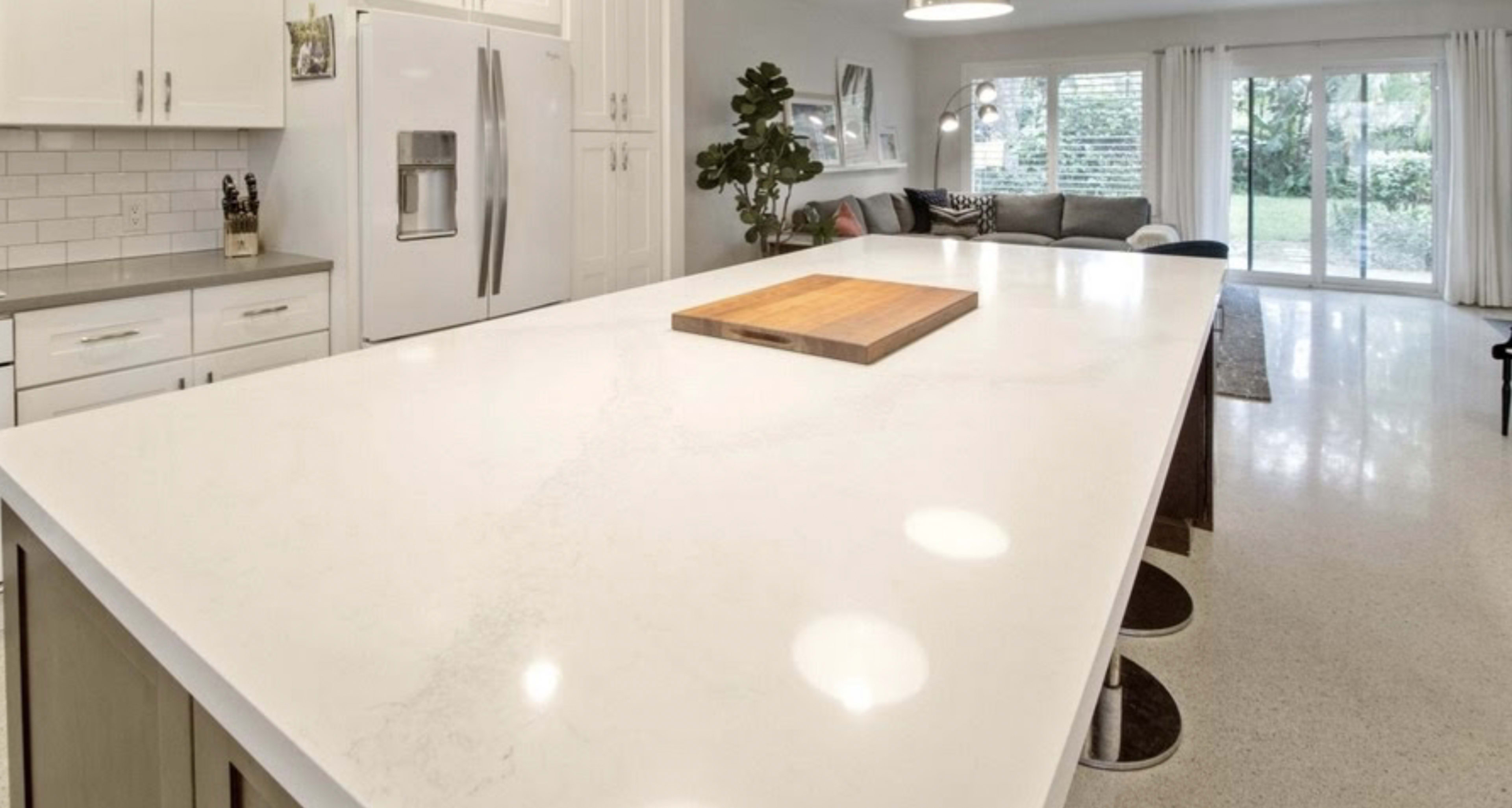 A modern kitchen features a large white countertop island with a wooden cutting board at its center, overlooking a cozy living area and a view of the outdoor greenery.