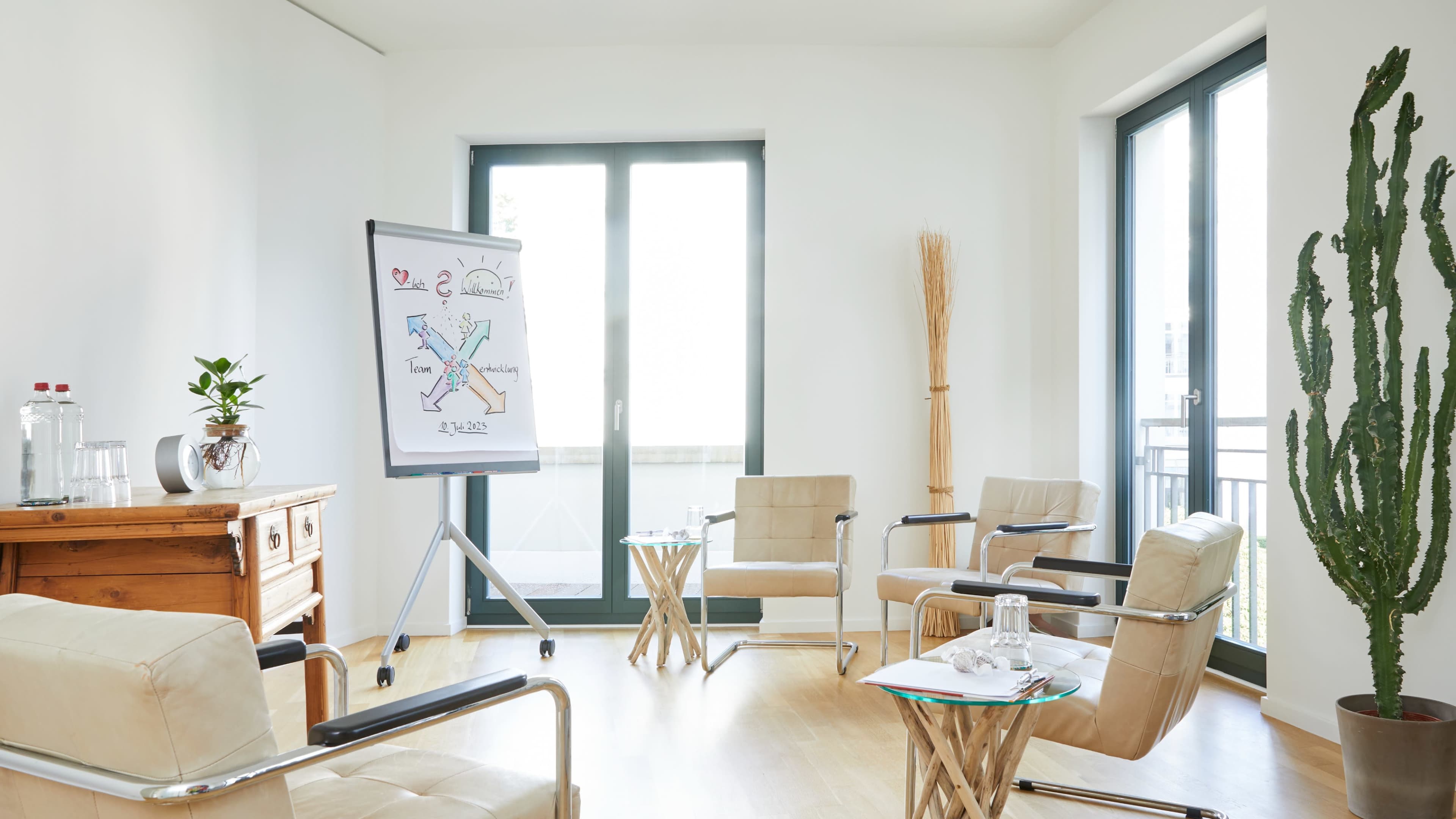 A well-lit meeting room features a whiteboard with diagrams, four beige chairs arranged around a wooden table, and a large cactus in the corner.