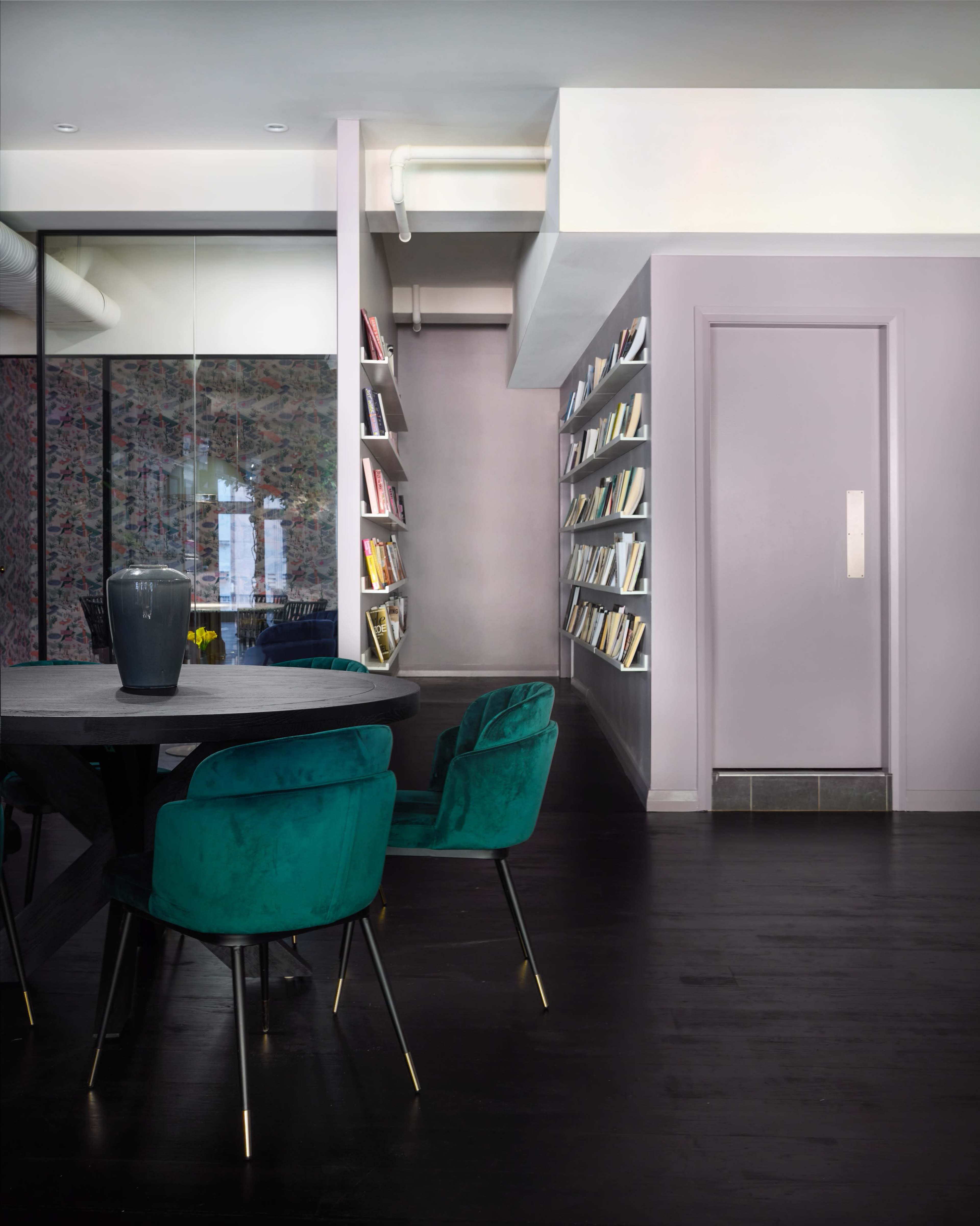 A modern interior features a round table with teal chairs and a wall-mounted bookshelf filled with books, alongside a door and a glass partition.