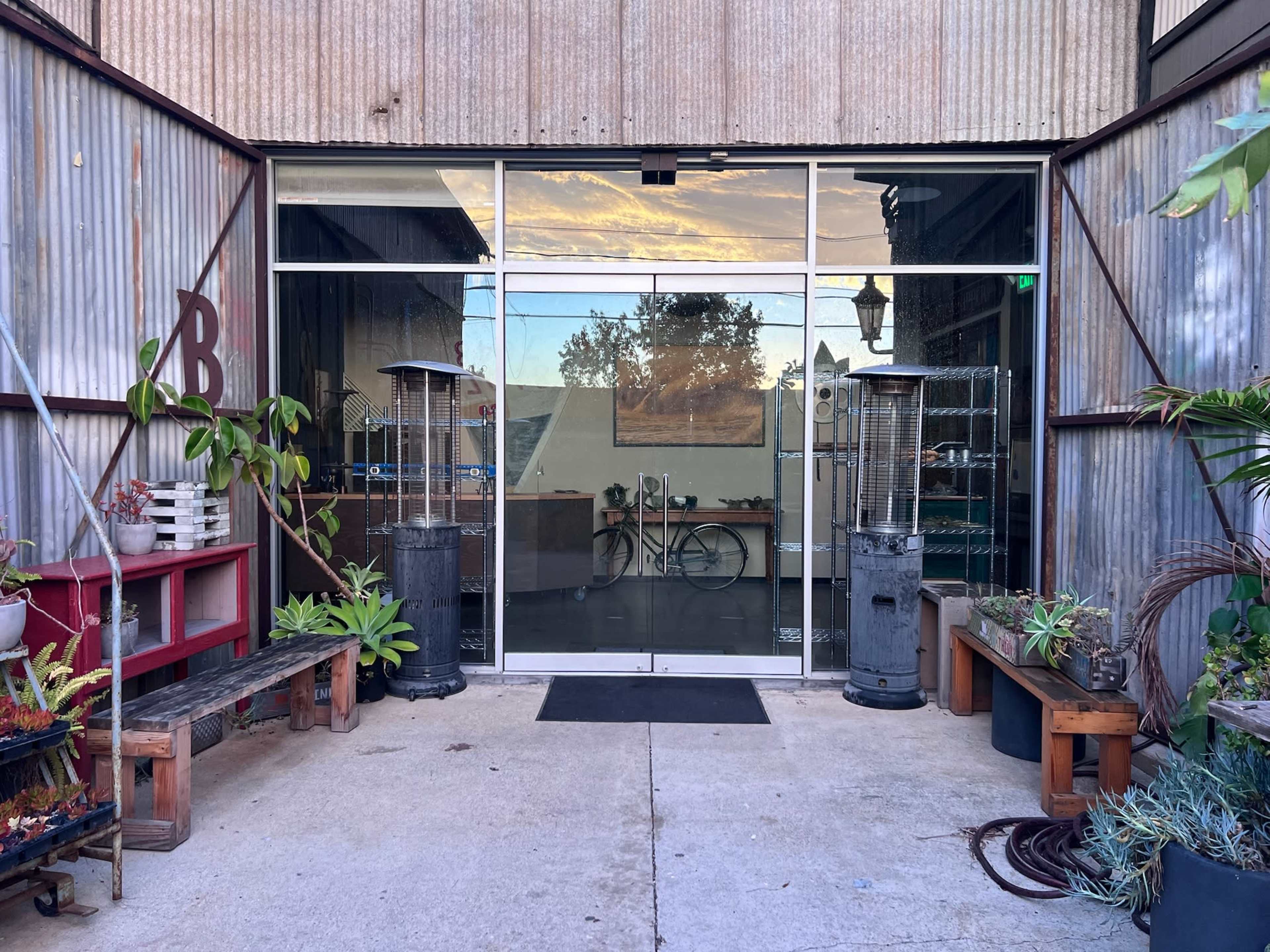 The image shows the entrance of a building with large glass doors, flanked by outdoor seating areas with potted plants and two patio heaters.
