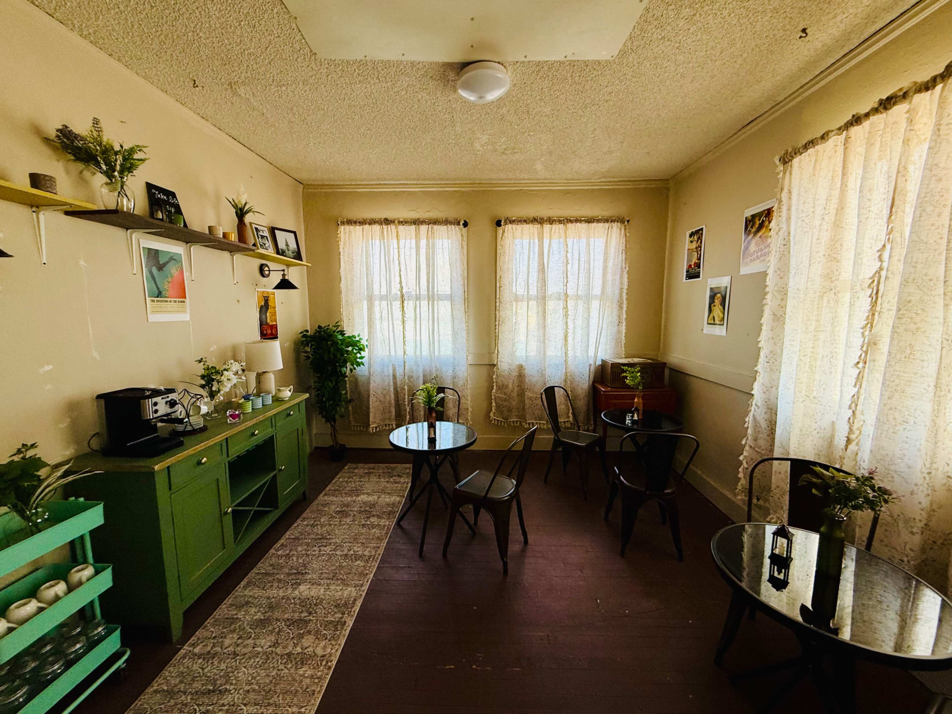 The room features a green console table, two small round tables with chairs, and large windows draped with sheer curtains, all arranged in a cozy setting.