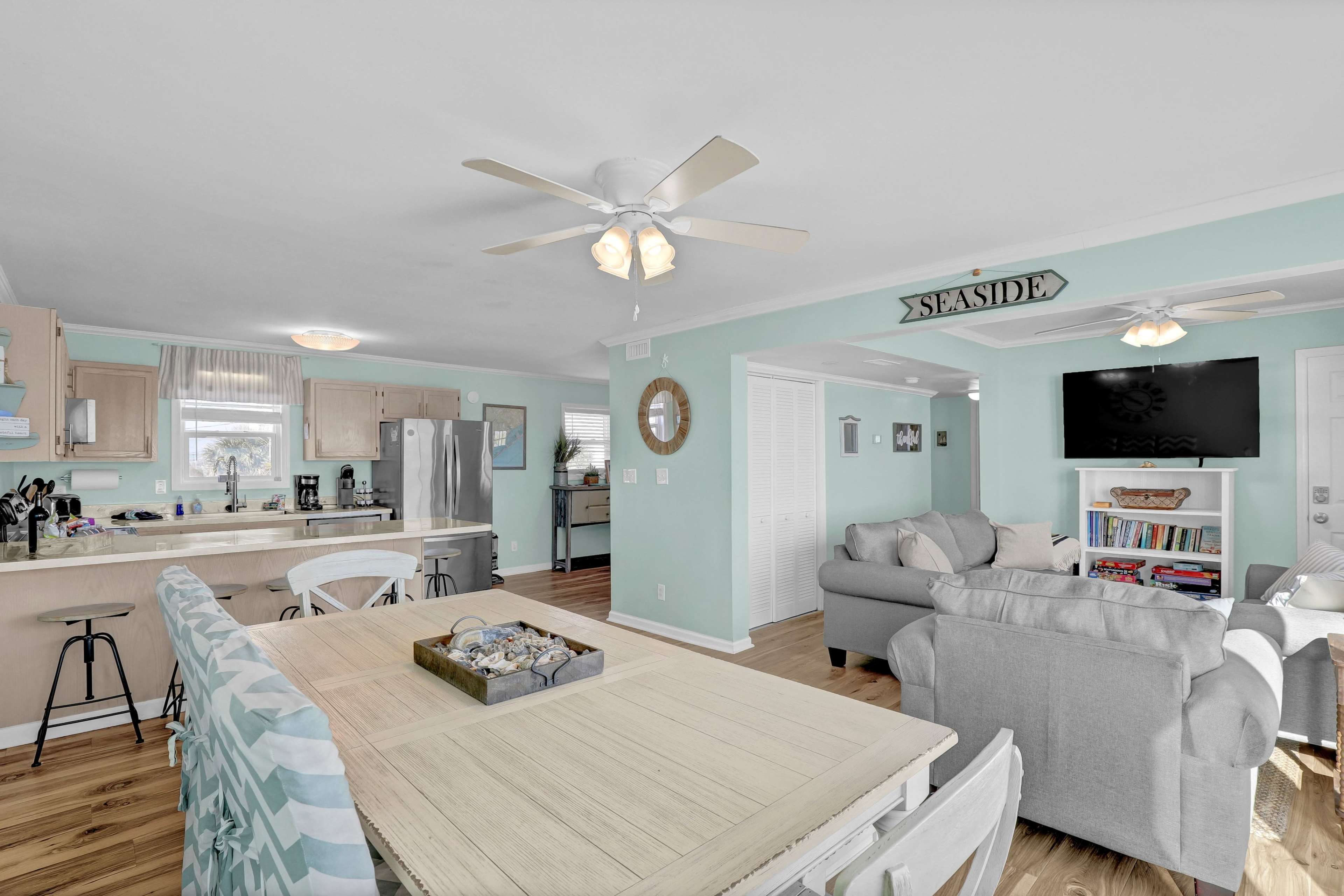 The image shows an open-concept living space featuring a dining area with a light-colored table, a kitchen with stainless steel appliances, and a cozy seating area with a TV.