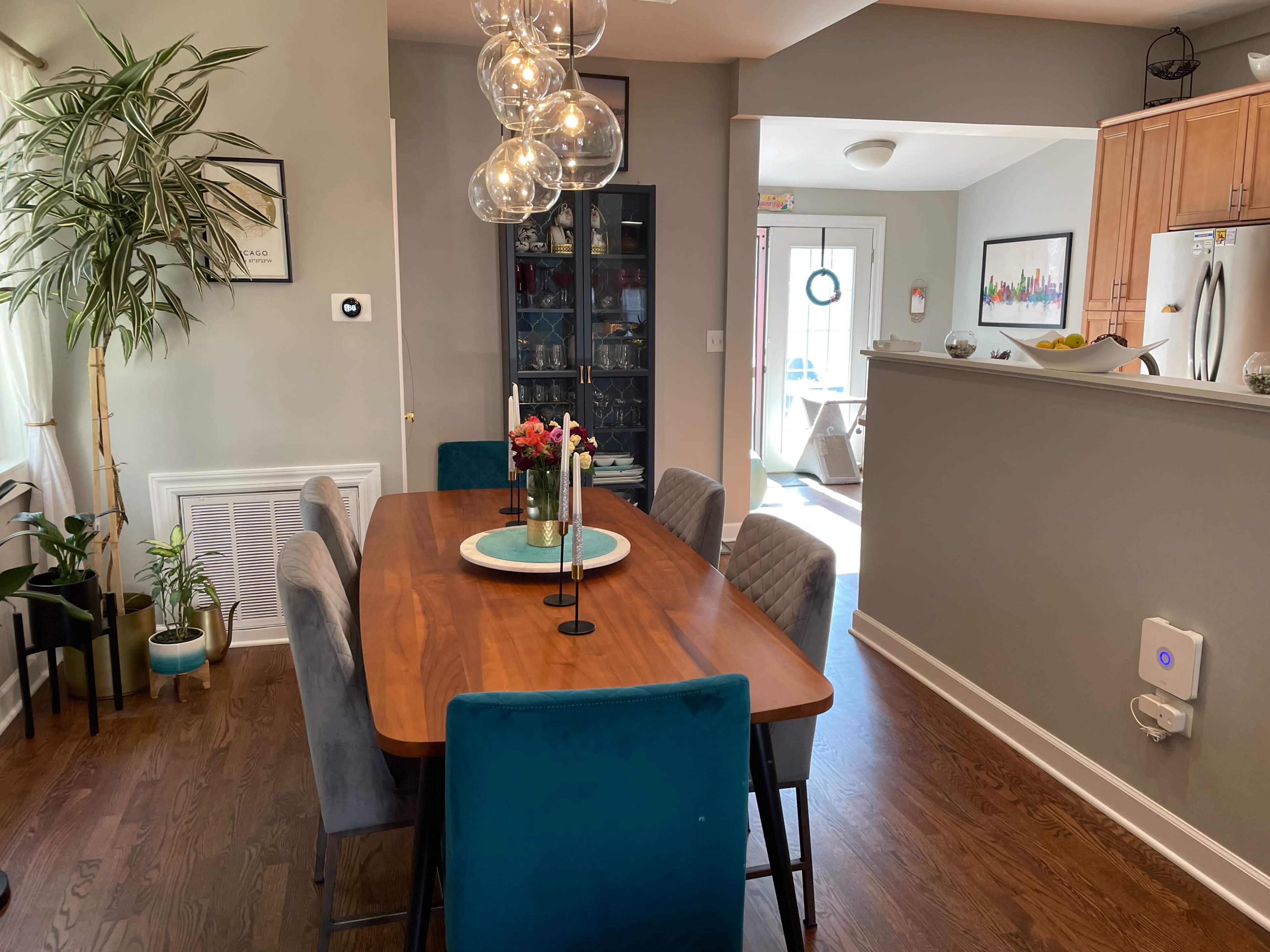 A wooden dining table with gray and teal chairs is positioned in a well-lit dining area that features a large window and a potted plant.