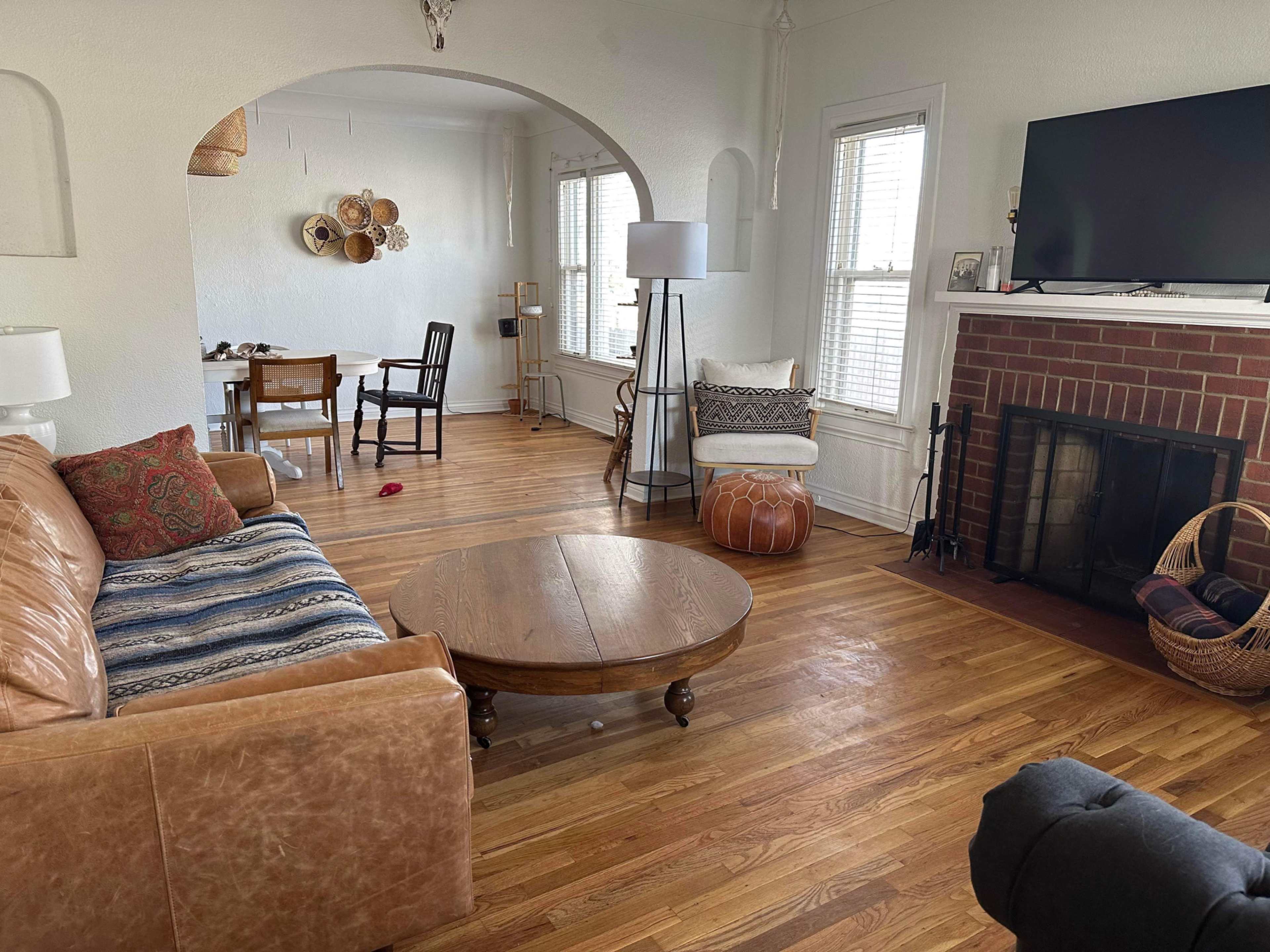 A living room with a brown leather sofa, a round wooden coffee table, a television mounted on the wall, and a fireplace surrounded by wooden flooring and various furnishings.