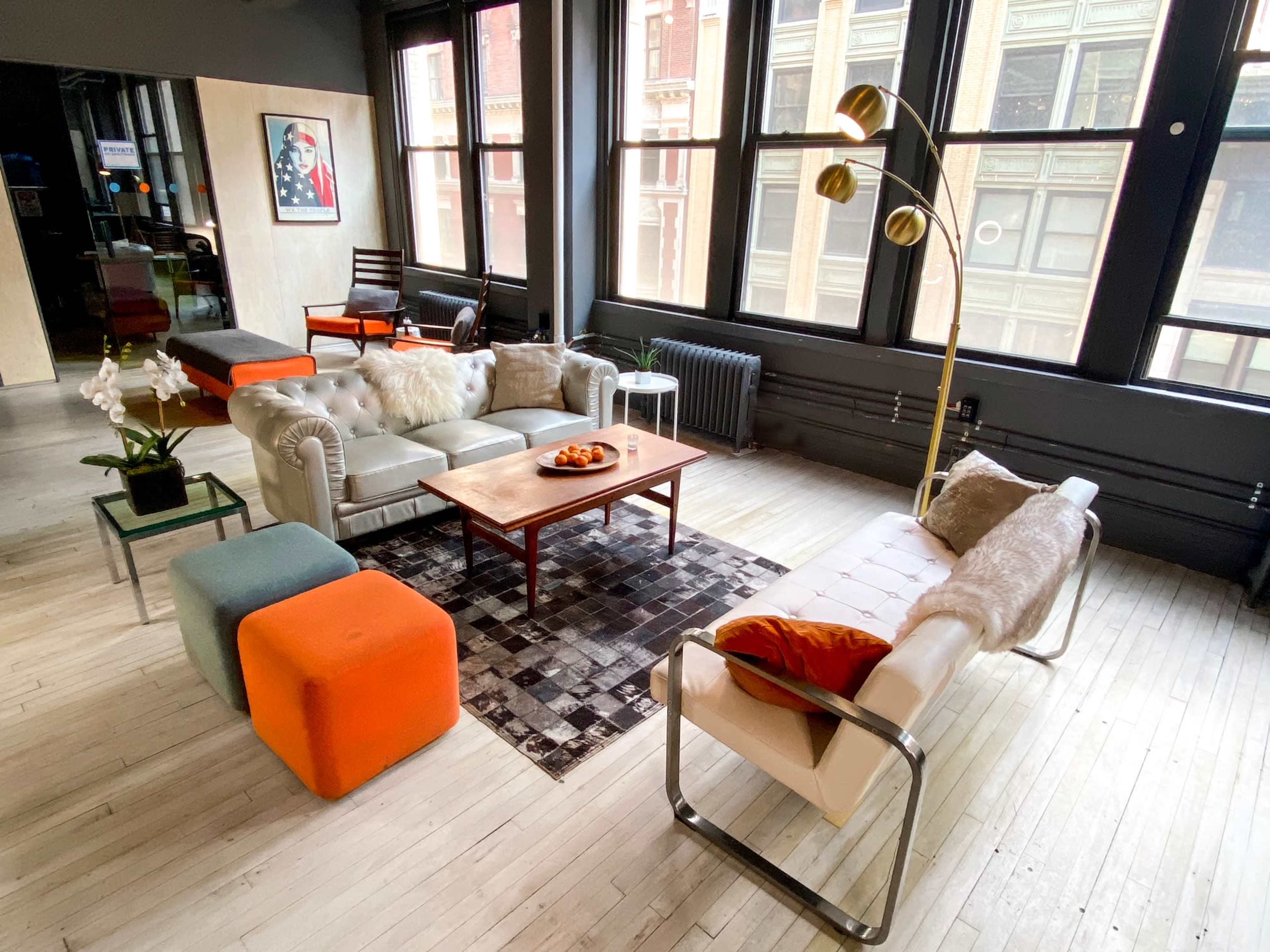 A modern living room with a mix of furniture, including a leather couch, a coffee table, and colorful ottomans, set in a spacious, well-lit area with large windows.