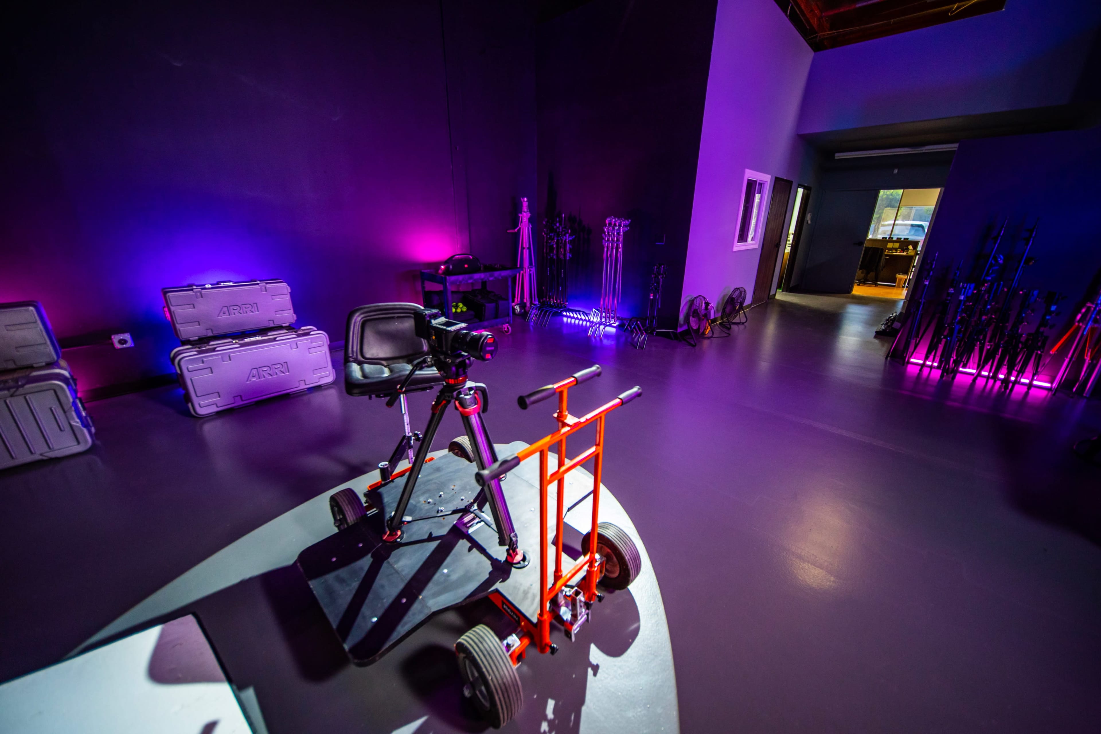 A spacious studio with a black and purple backdrop, featuring a wheeled camera stabilizer and various filmmaking equipment stored in cases along the walls.