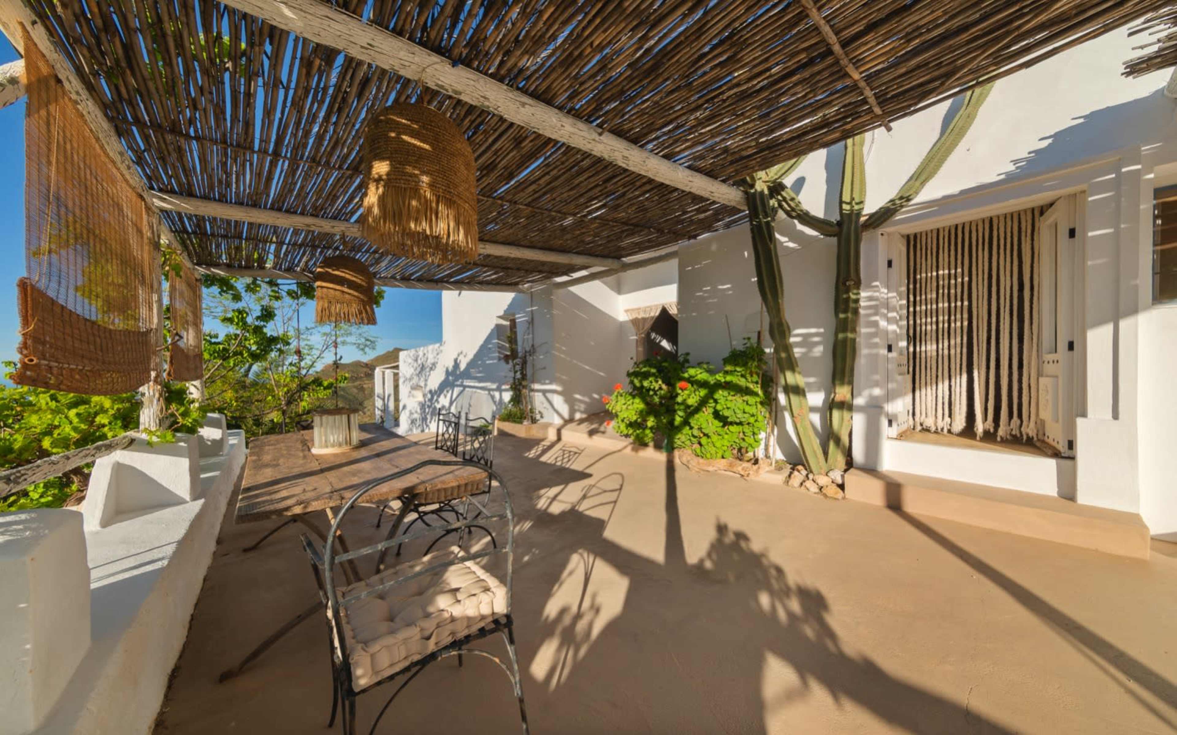 The image shows a sunlit outdoor patio with a wooden table, chairs, and a thatched roof, surrounded by greenery and white walls.