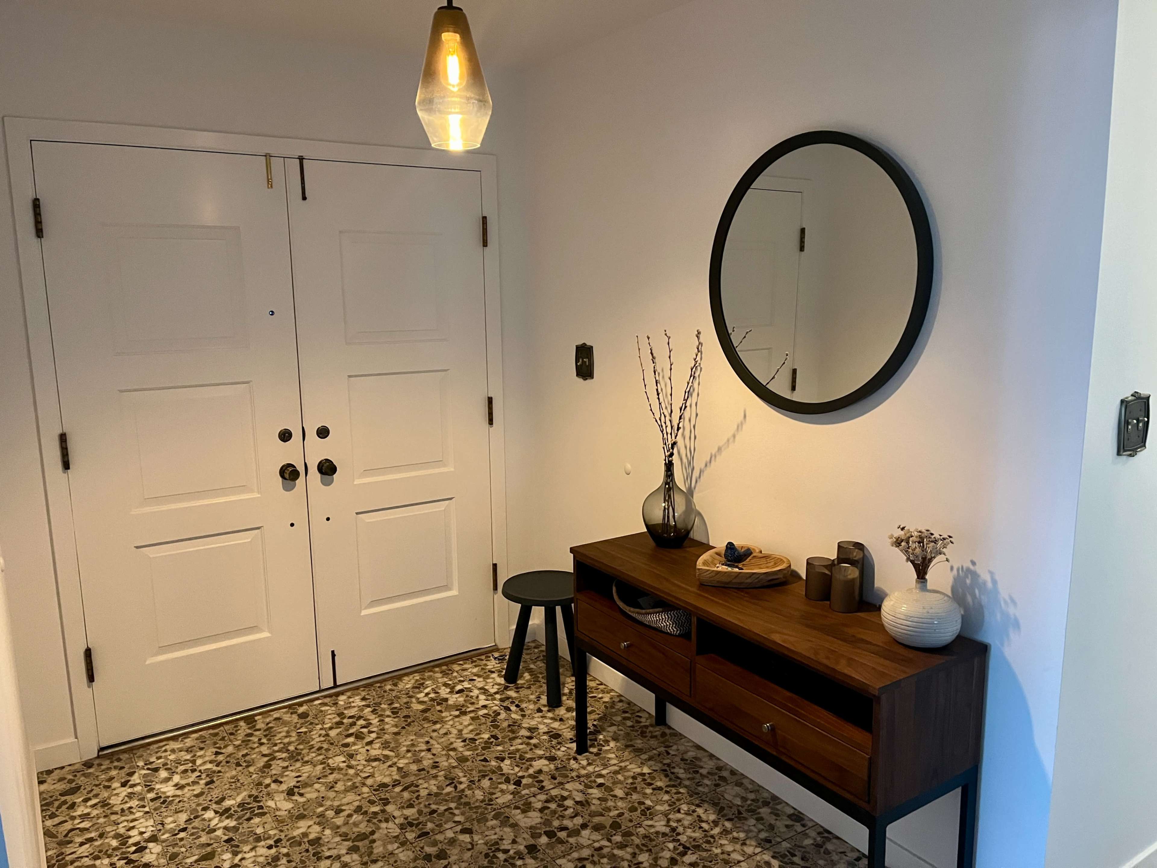 An entryway featuring two doors, a round mirror above a wooden console table, and a stool, with decorative items and a patterned floor.