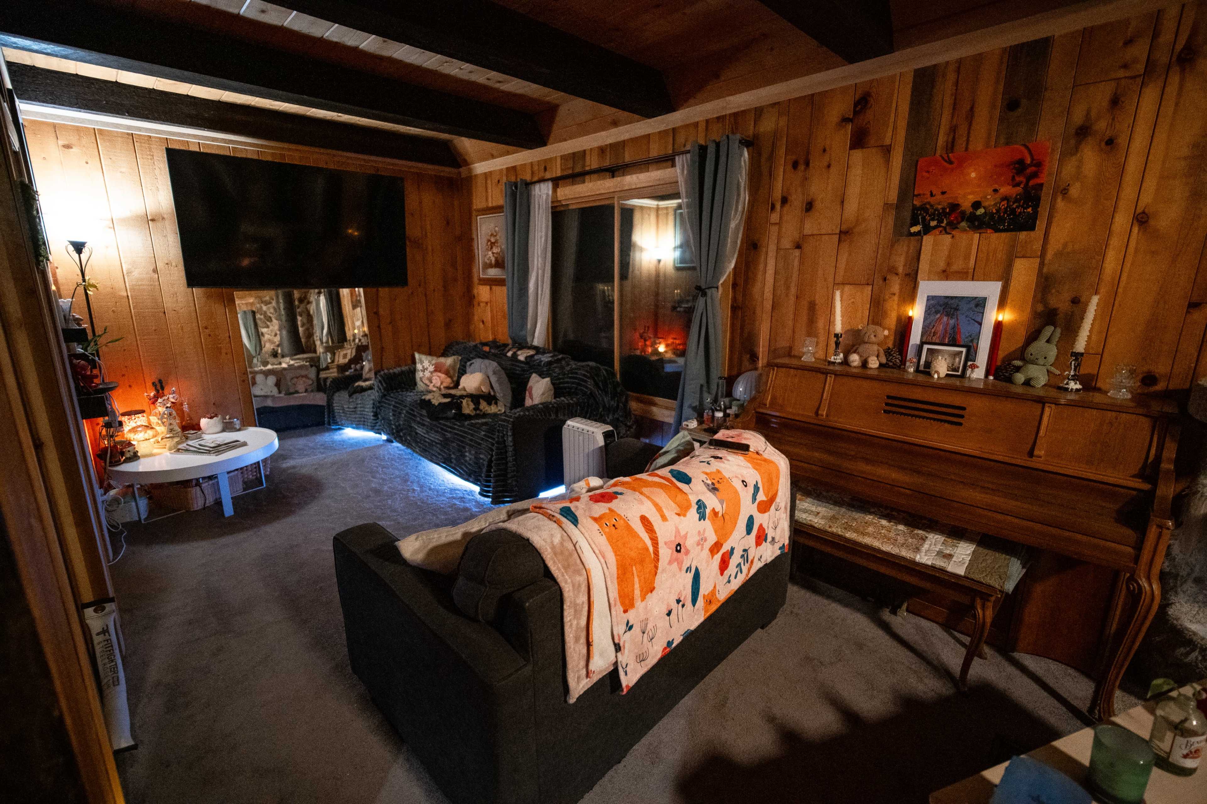 The image shows a cozy living room with wooden walls, a dark couch with a colorful blanket draped over it, a piano, and a large television mounted on the wall.